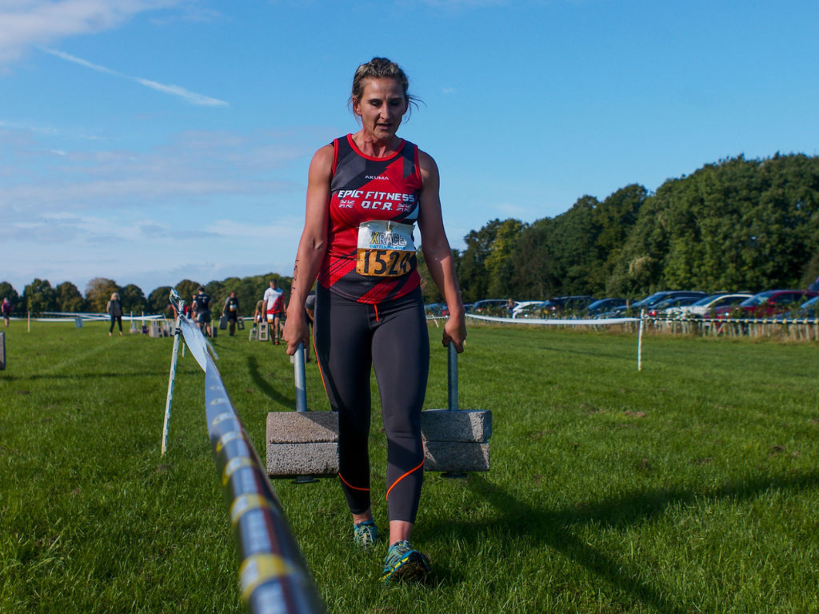 A person wearing a red athletic shirt and black leggings carries concrete blocks down a grassy outdoor course. The individual appears focused and determined. Other participants and spectators are visible in the background. The sky is clear and the day is bright.