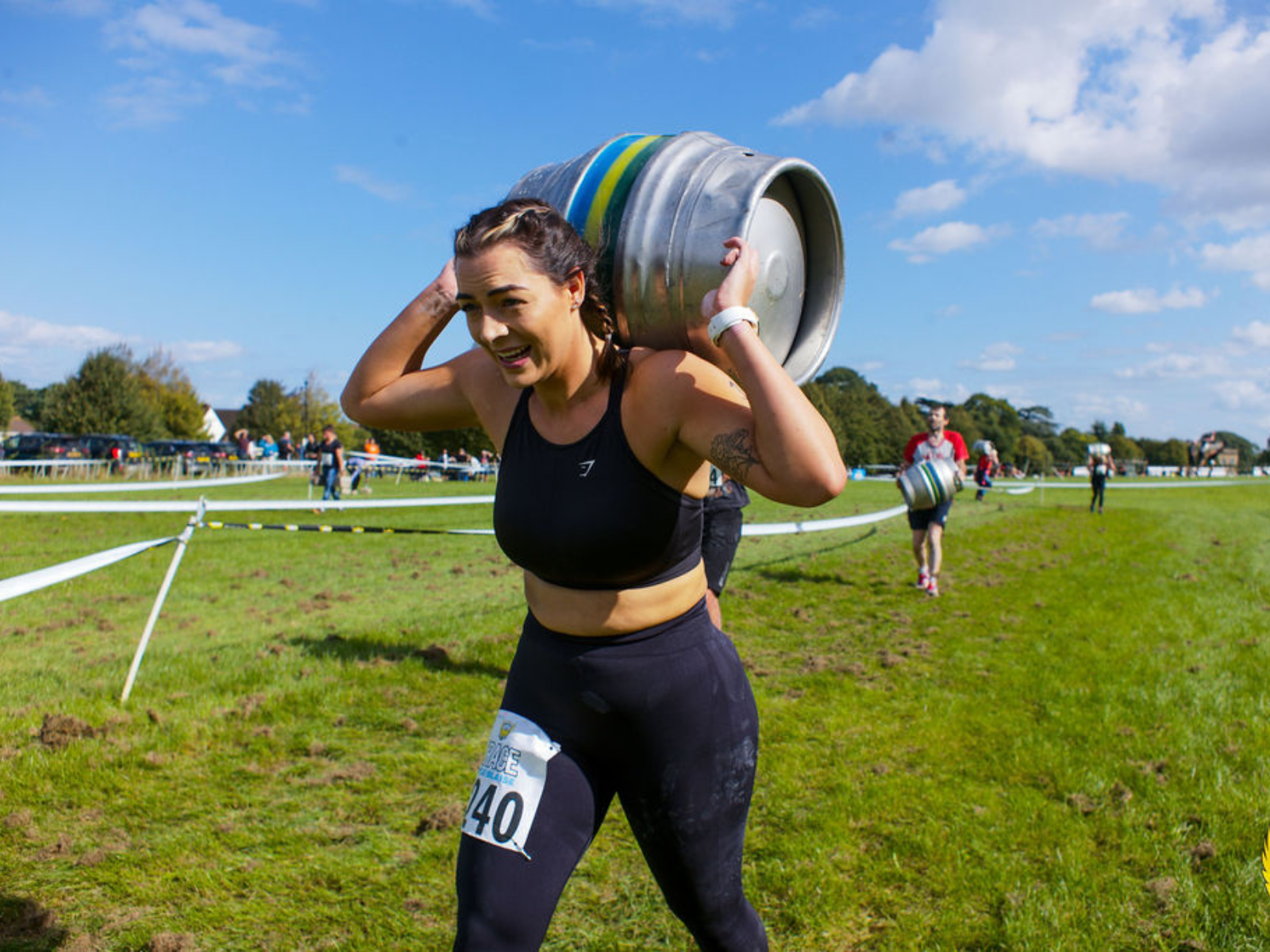A woman, wearing a black sports bra and leggings, carries a large metal keg on her shoulders while running on a grassy racecourse. Other participants and spectators are visible in the background. She has the number 40 pinned to her leggings.