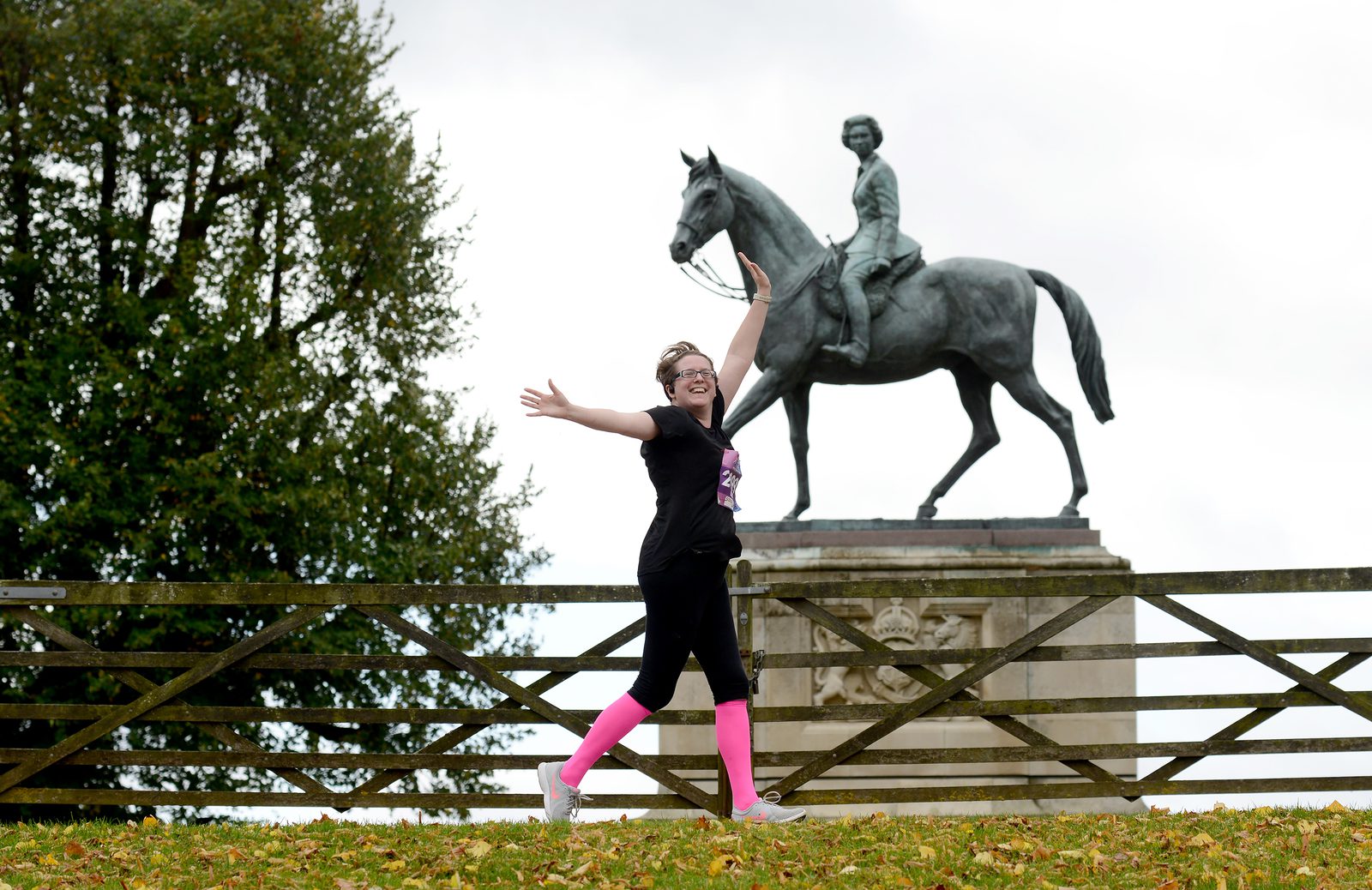 A person with short hair, wearing a black t-shirt, black leggings, and bright pink leg warmers joyfully poses with arms outstretched near a horse statue. The background includes a wooden fence and green trees. The sky is overcast.