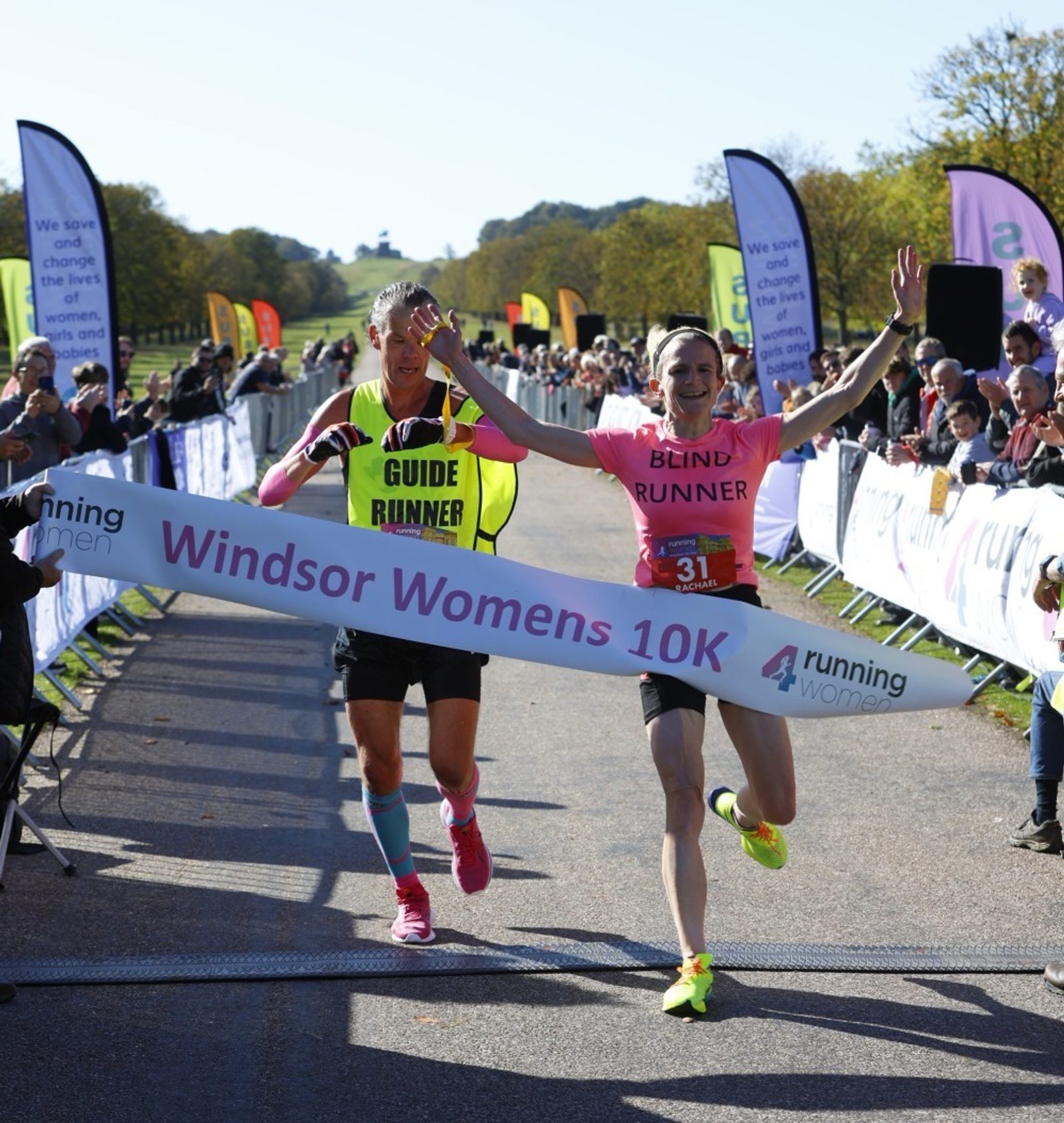 A blind runner crosses the finish line of the Windsor Women's 10K with the help of a guide. Both are smiling and cheering. Spectators and colorful flags line the sides of the sunlit race path.