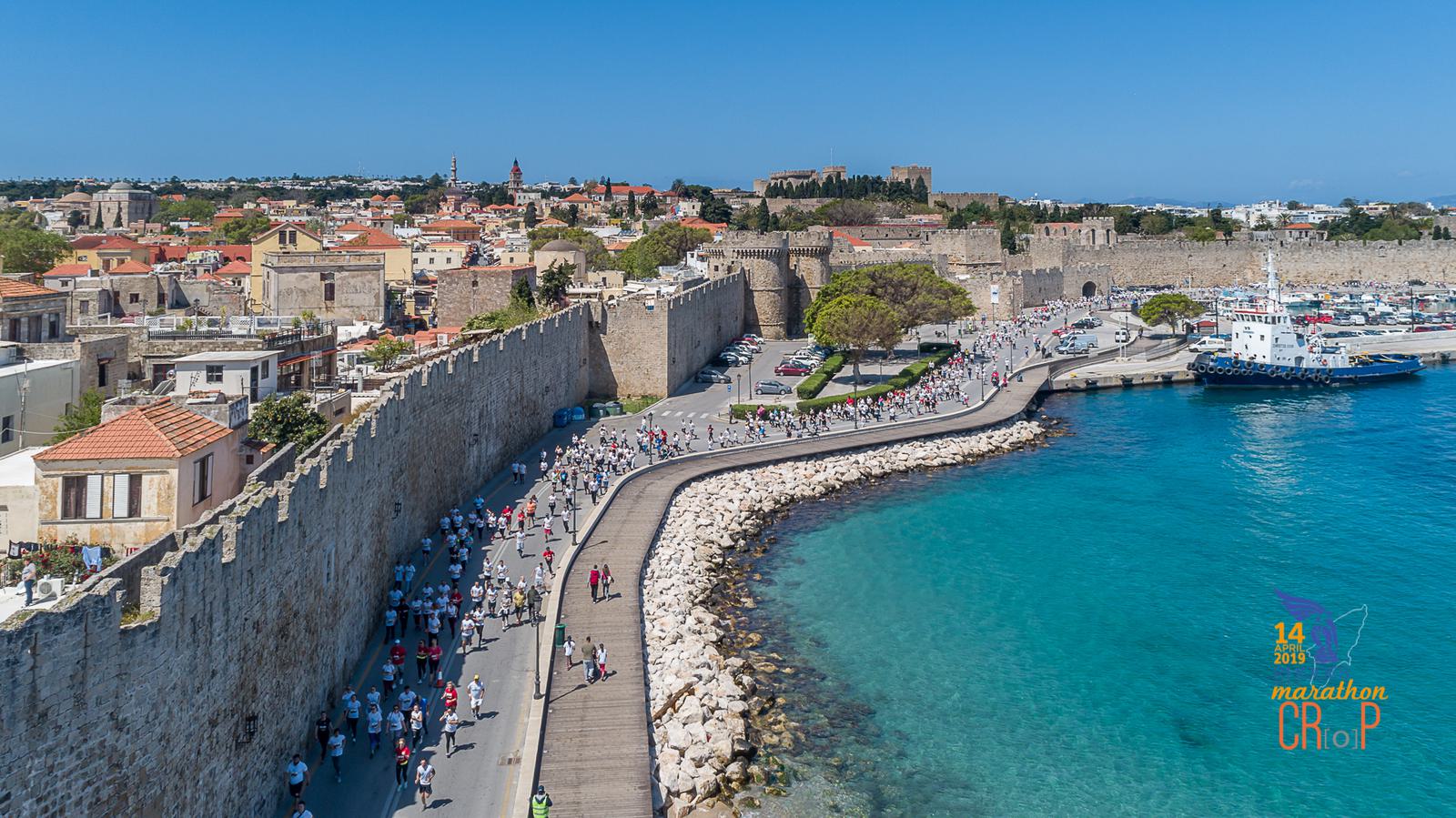 A large group of people walk along a coastal path next to ancient city walls, with turquoise water on one side and historic buildings on the other. The scene is vibrant with participants likely in a marathon or public event. A ship is docked nearby.