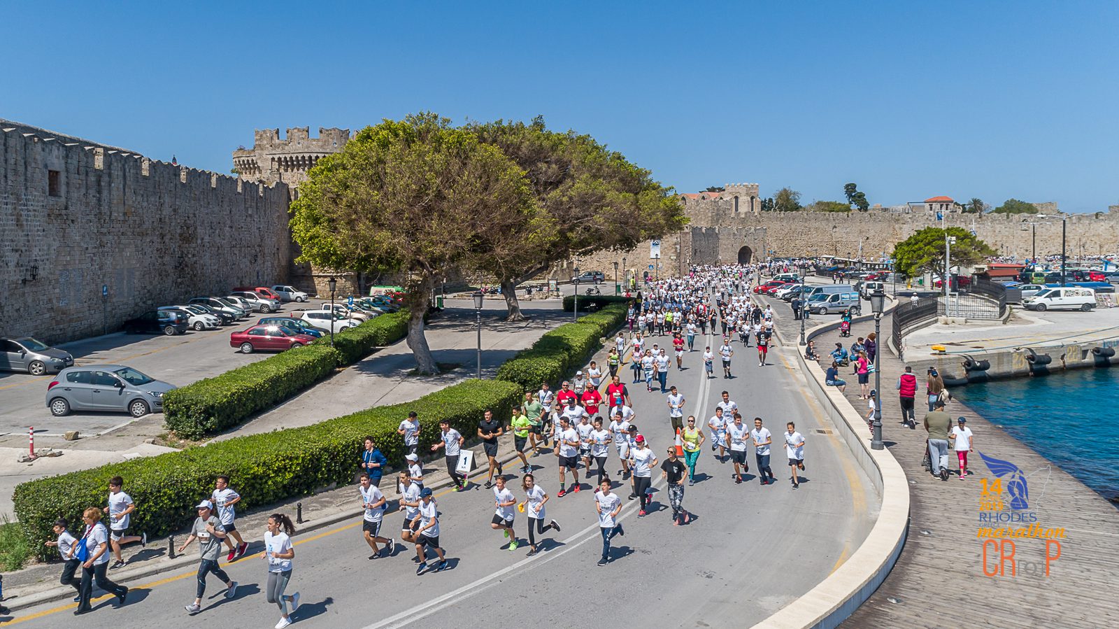 An aerial view shows a crowd of people participating in a marathon on a sunny day. They are running on a road next to water, with parked cars and a large stone fortress visible in the background. Trees line parts of the road, and participants are wearing event t-shirts.