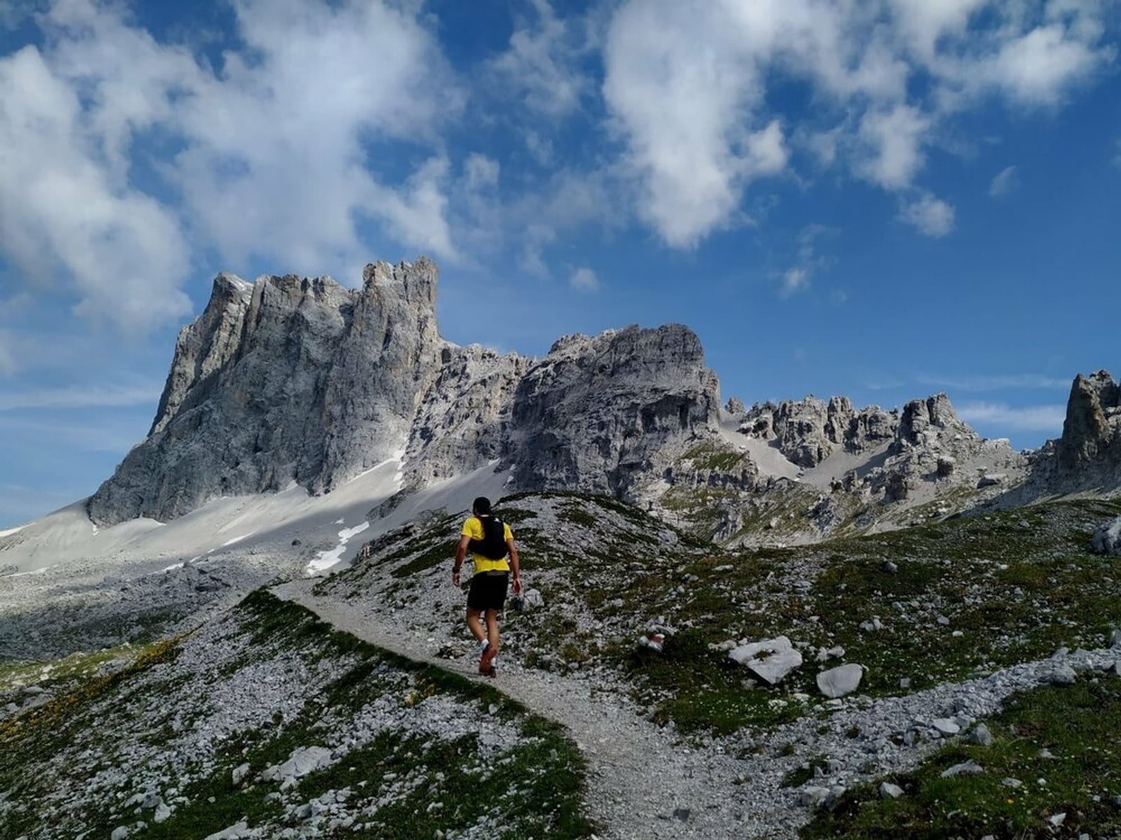 A hiker wearing a yellow backpack and black shorts walks along a rocky trail toward a towering, craggy mountain under a blue sky with scattered clouds. The mountain's rugged terrain is contrasted by patches of green vegetation along the path.