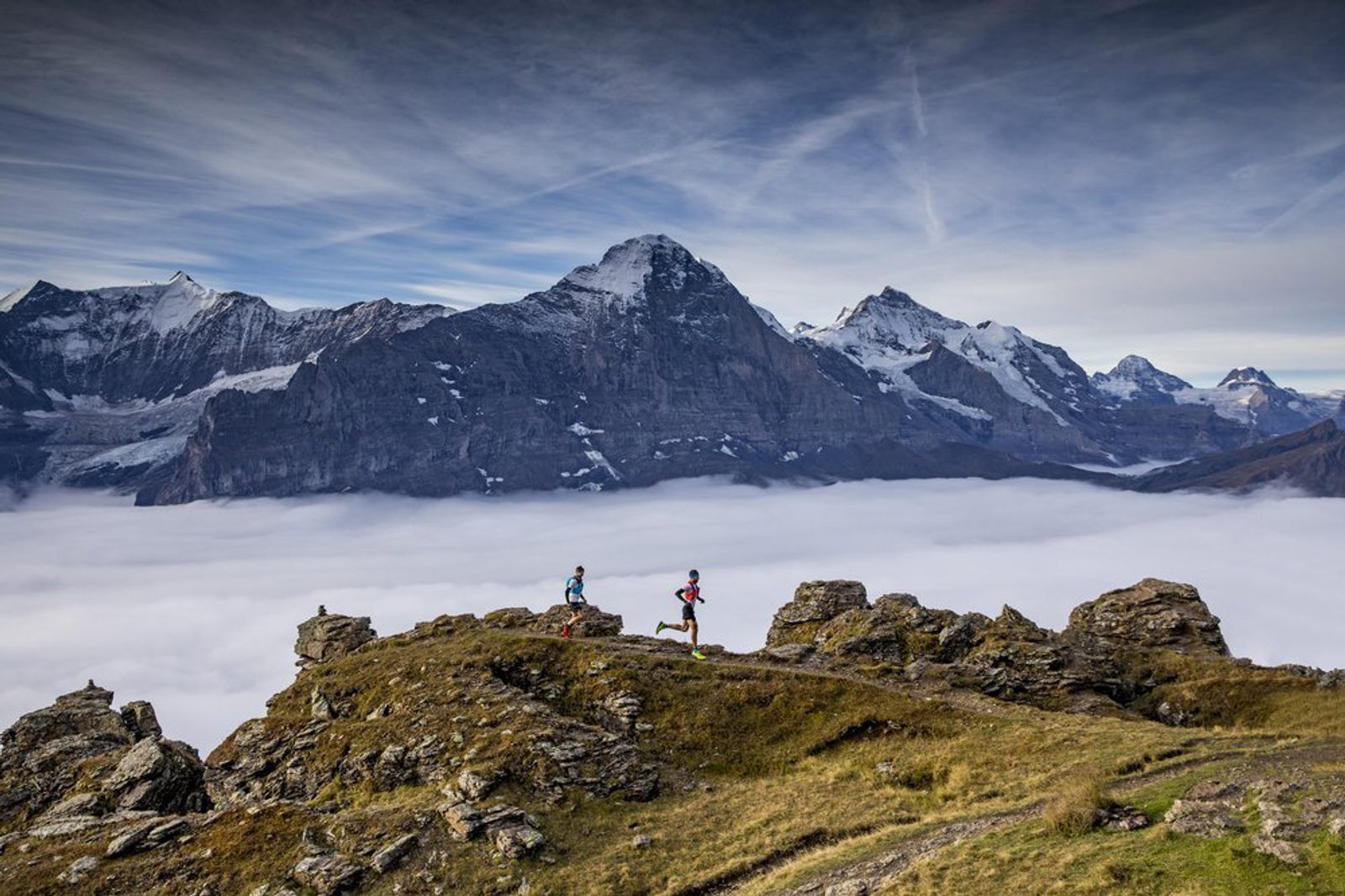 Two runners are traversing a rocky mountain ridge under a sky streaked with thin clouds. Snow-capped peaks rise majestically in the background, while a blanket of clouds fills the valley below, creating a stunning, high-altitude landscape.