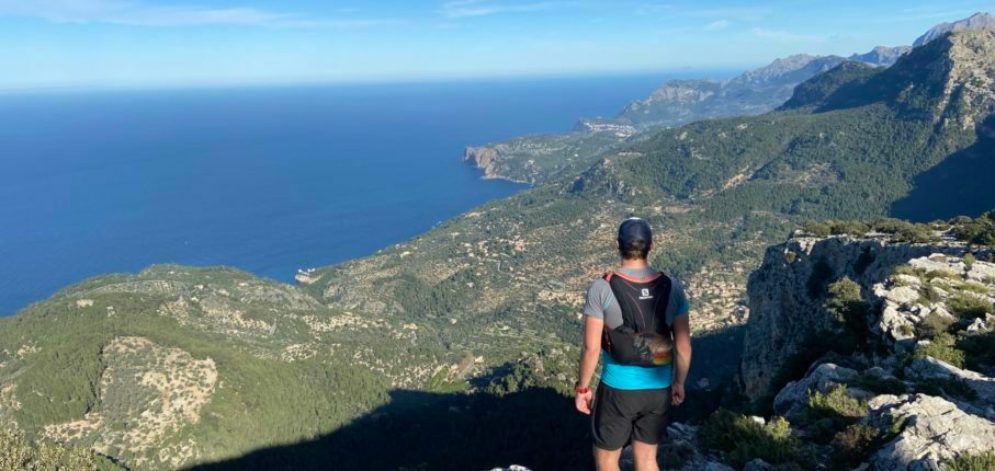 A person wearing hiking gear stands on a rocky ledge, overlooking a vast expanse of green hills and the blue sea under a clear sky. The view includes a rugged coastline, distant mountains, and scattered settlements.