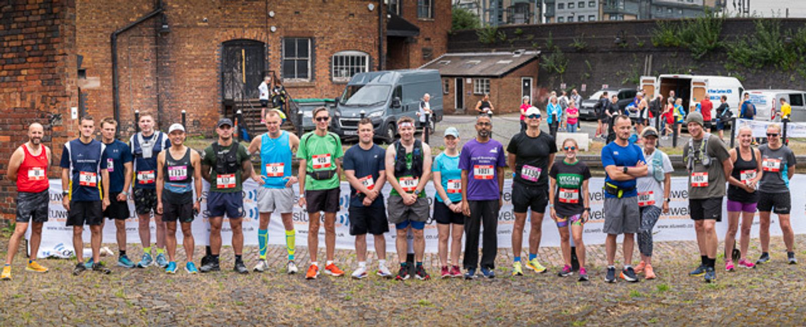 A group of runners in athletic gear stand side by side in front of an industrial-style backdrop, preparing for a race. Each participant wears a bib number, and some are smiling at the camera. Various spectators and event staff can be seen in the background.