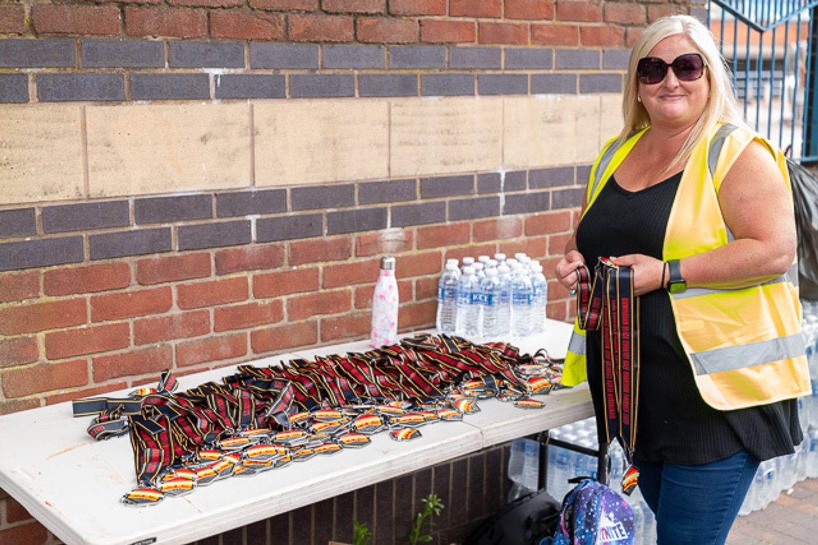 A woman wearing a high-visibility vest stands next to a table covered with medals and water bottles. She is smiling and holding several medals. The background features a brick wall and a partial view of a staircase railing.