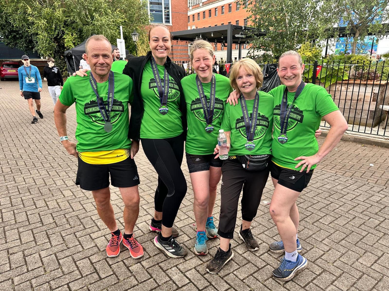 Five people in matching green shirts and medals smile together outdoors after a race. They stand on a brick walkway, some with hands on hips, appearing happy and accomplished. Trees and buildings are visible in the background.