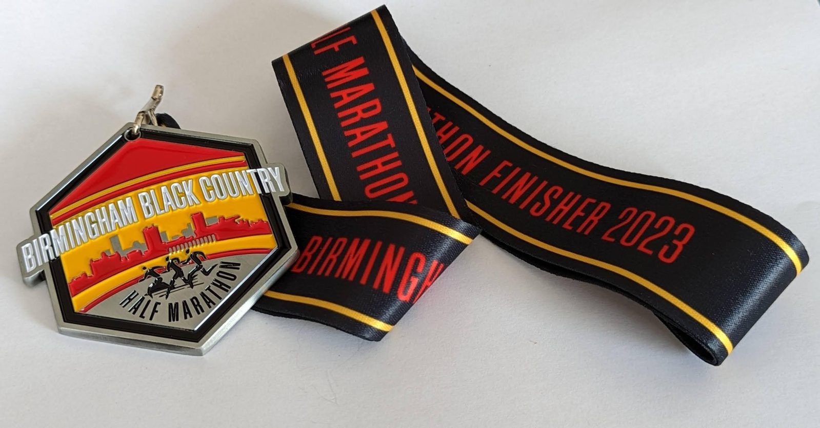 A medal with a hexagonal shape and a ribbon. The medal, labeled "Birmingham Black Country Half Marathon," features a skyline and runners. The ribbon is black with red and yellow accents, reading "Half Marathon Finisher 2023" and "Birmingham".