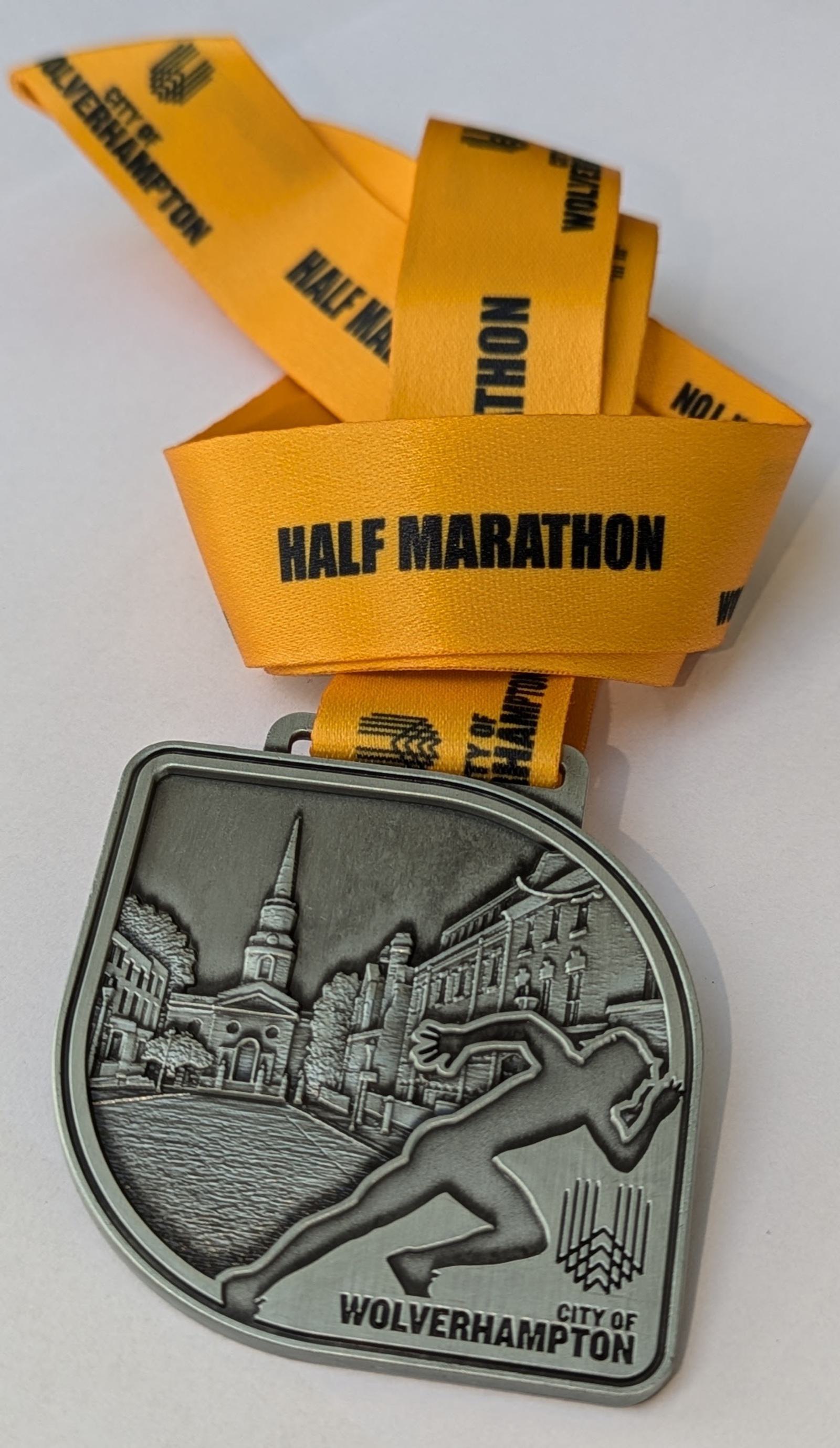 A silver medallion with a raised design of a running figure and buildings, hanging from an orange ribbon labeled "HALF MARATHON" and "CITY OF WOLVERHAMPTON.
