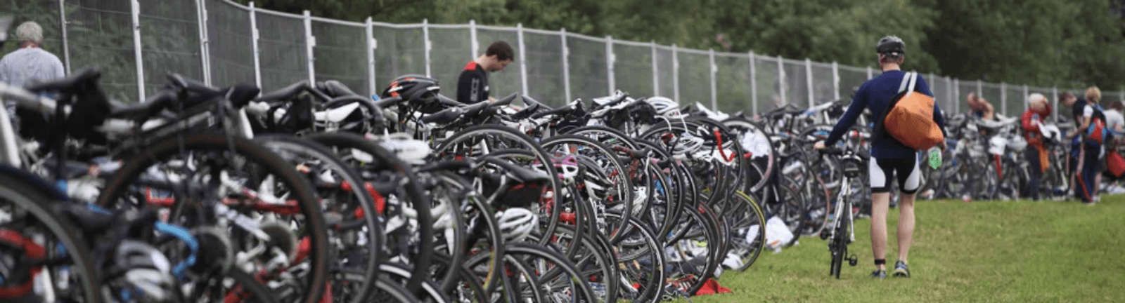 A group of people gather near a large number of parked bikes lined up along a fenced area. One person walks away carrying an orange bag and wearing a helmet while others can be seen in the background. The scene takes place outdoors with trees in the distance.