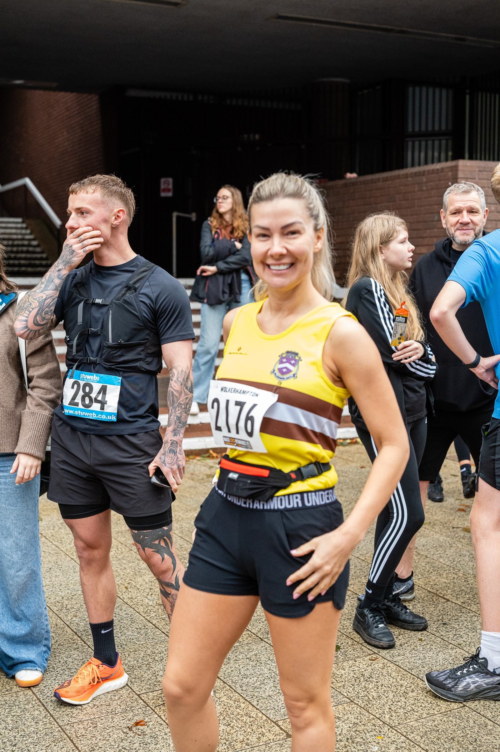 A woman wearing a yellow and brown running uniform with the number 2176 smiles and poses for the camera. A man with tattoos, wearing a black athletic outfit and the number 284, stands nearby, looking to the side. Other people are in the background.