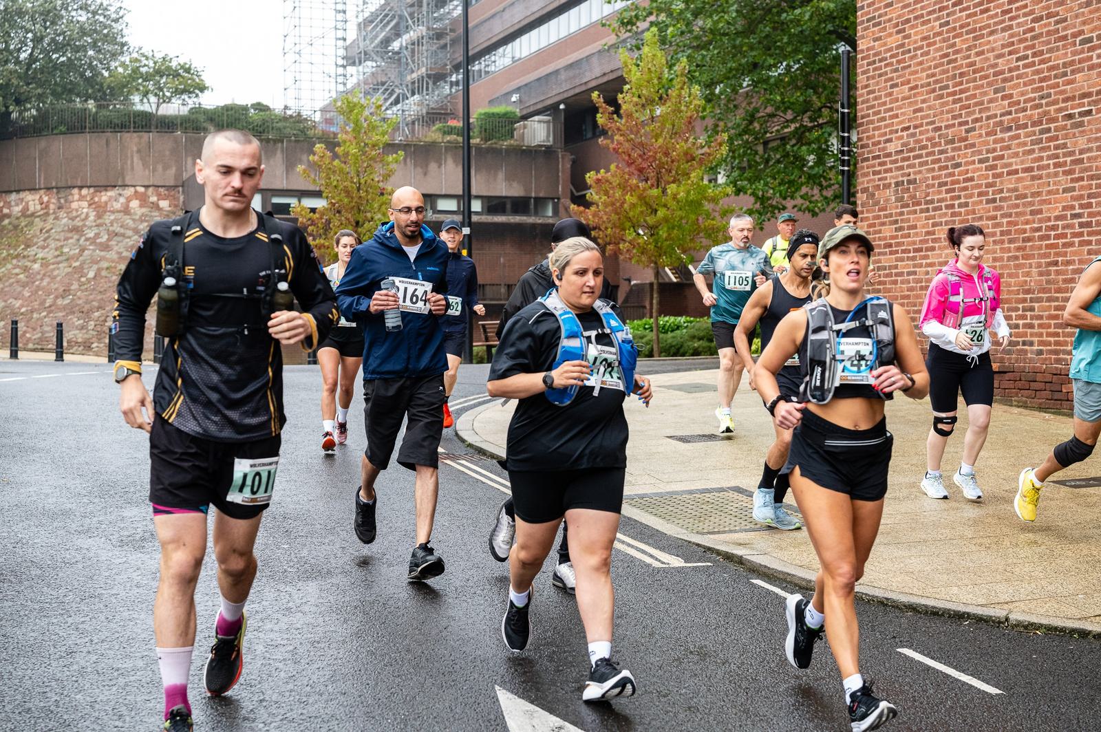 A group of runners, including men and women wearing numbered bibs, run on a wet street. The participants are in athletic gear with some wearing hydration packs. A brick building and trees are in the background. The sky is overcast.