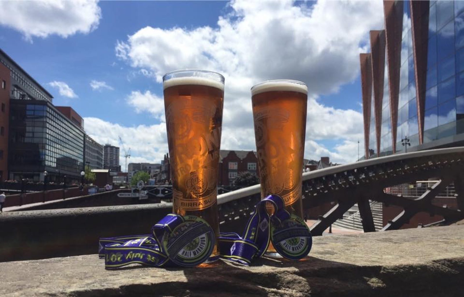 Two tall glasses of amber beer stand on a stone surface, each adorned with a blue and gold medal draped around its base. The background features a cityscape with modern buildings, a bridge, and a partly cloudy blue sky.