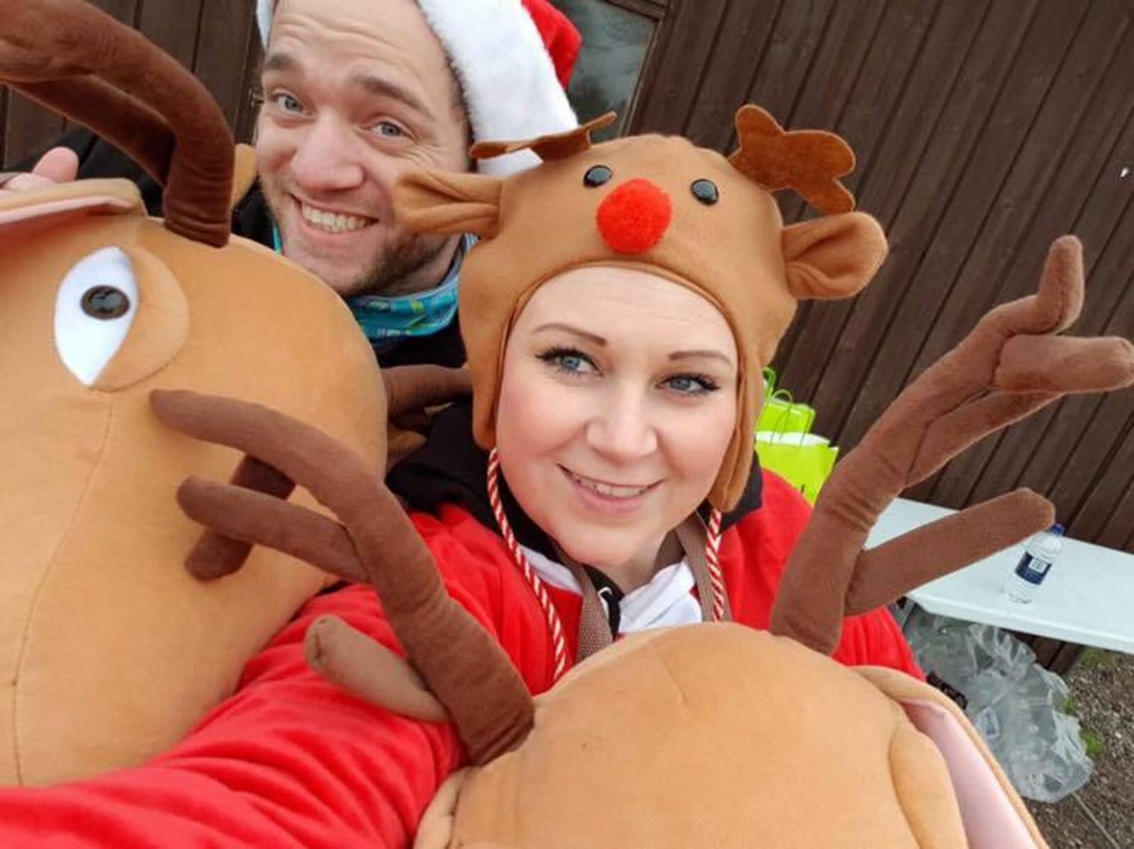 A man and woman dressed in festive costumes smile at the camera. The man is wearing a Santa hat and reindeer antlers, while the woman is wearing a reindeer hat with a red nose and antlers. They appear to be enjoying a holiday celebration.