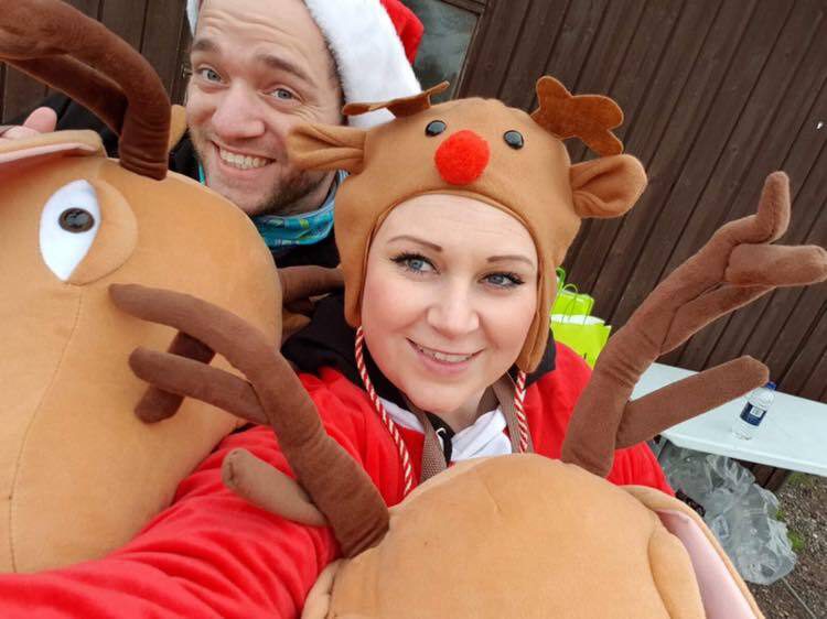 A man and woman dressed in festive costumes smile at the camera. The man is wearing a Santa hat and reindeer antlers, while the woman is wearing a reindeer hat with a red nose and antlers. They appear to be enjoying a holiday celebration.