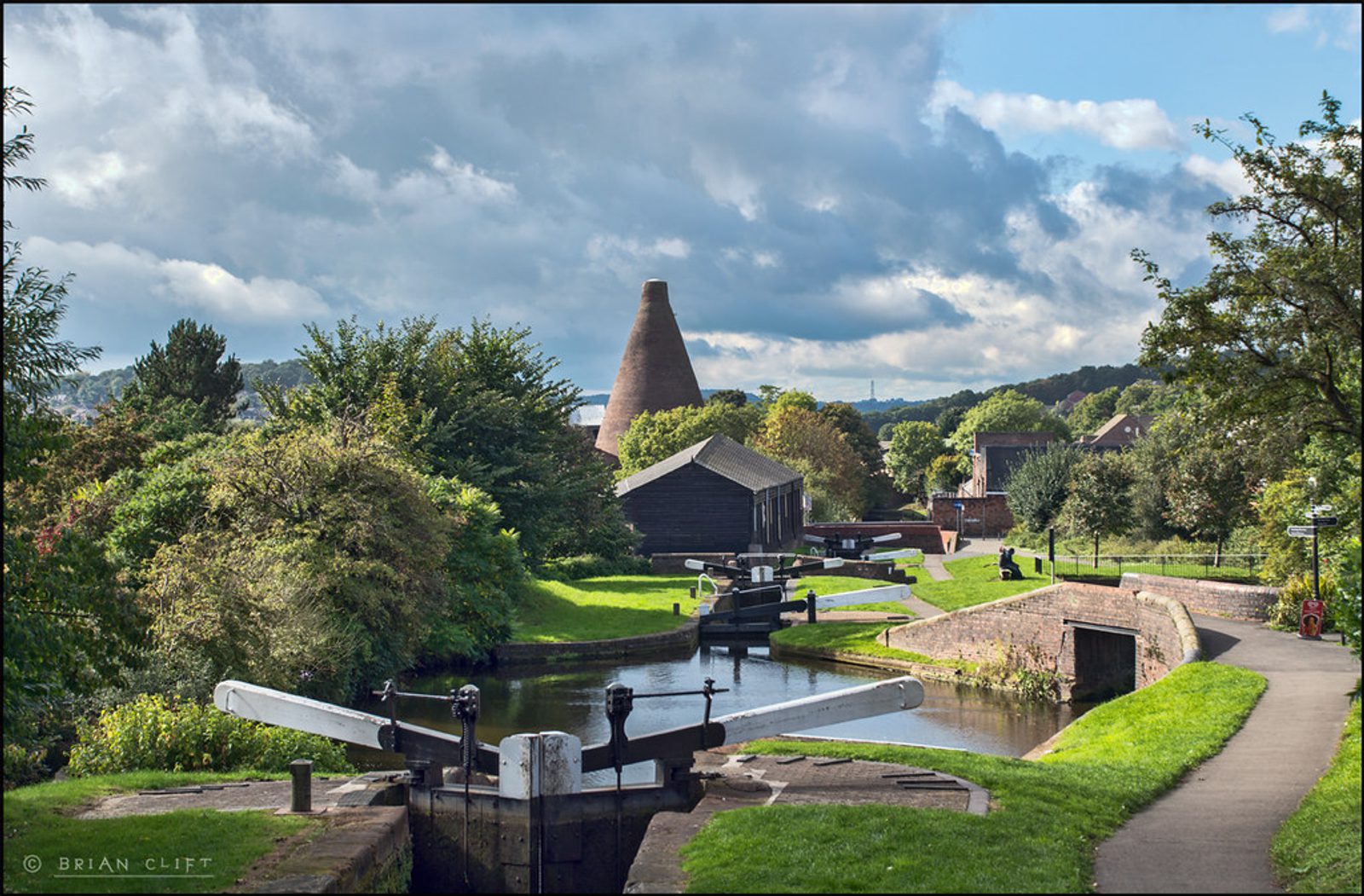 This image depicts a scenic canal with lock gates, bordered by lush greenery and trees. In the background, there are historic buildings. Among the structures is a prominent, unique-looking bottle kiln standing tall against a partly cloudy sky.