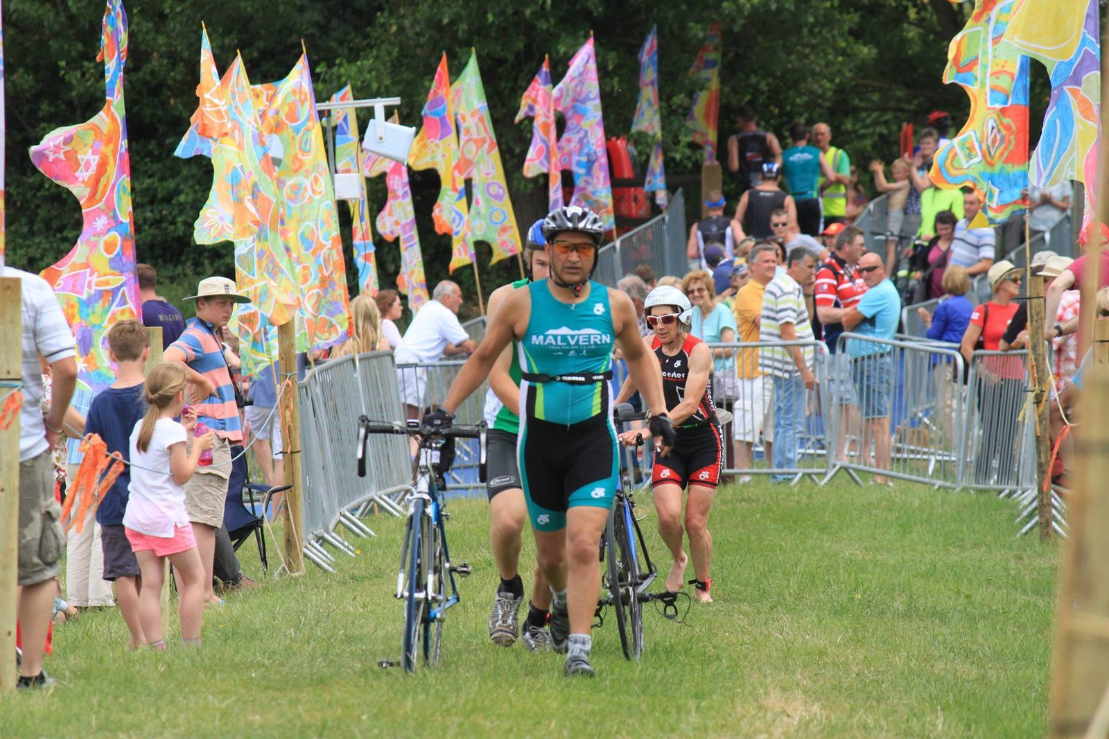Two athletes in triathlon gear walk their bicycles through a grassy course lined with vibrant, colorful flags. Spectators stand behind barriers, watching the event. The scene is lively with people of various ages, wearing casual summer clothing.