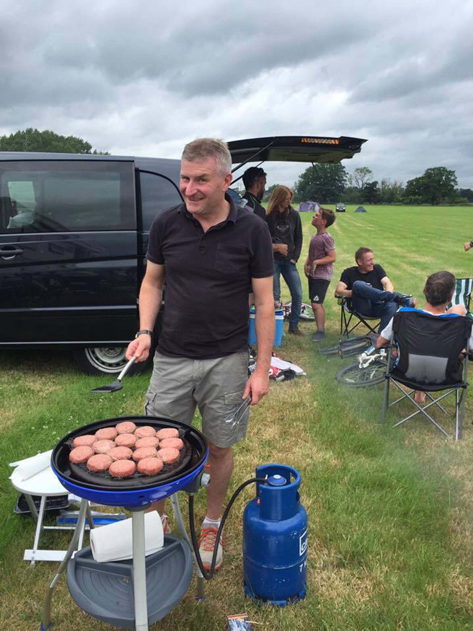 A man is grilling burgers on a portable grill in an open grassy area. He is wearing a black polo shirt and khaki shorts. Several people are seated and standing around nearby, and a black van is parked behind them. The sky is cloudy.