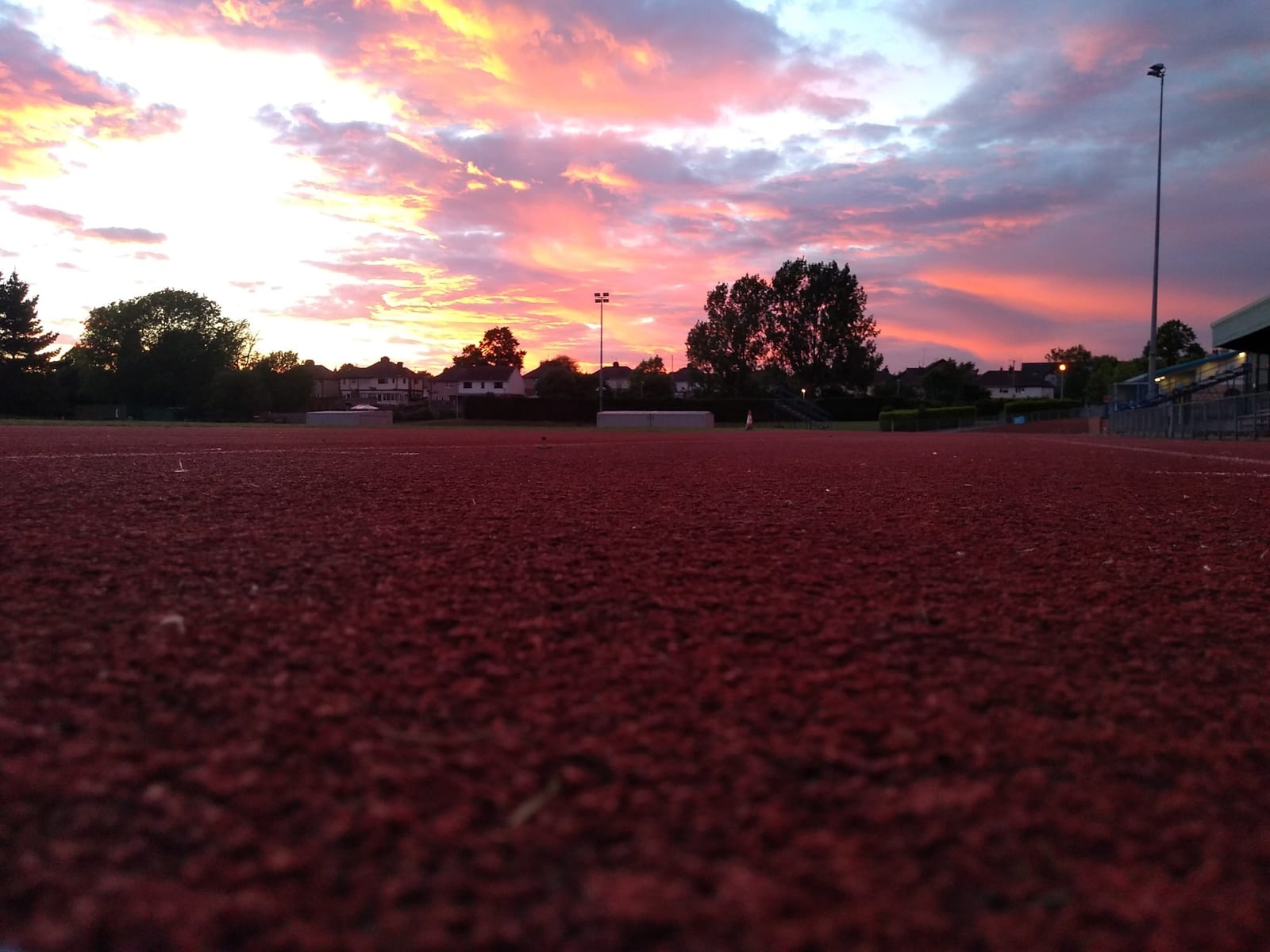 A track field at sunset, with a vibrant sky painted in hues of pink, orange, and purple. The sun is setting behind the silhouettes of trees and buildings in the background, casting a warm glow over the scene. The track's red surface is prominent in the foreground.