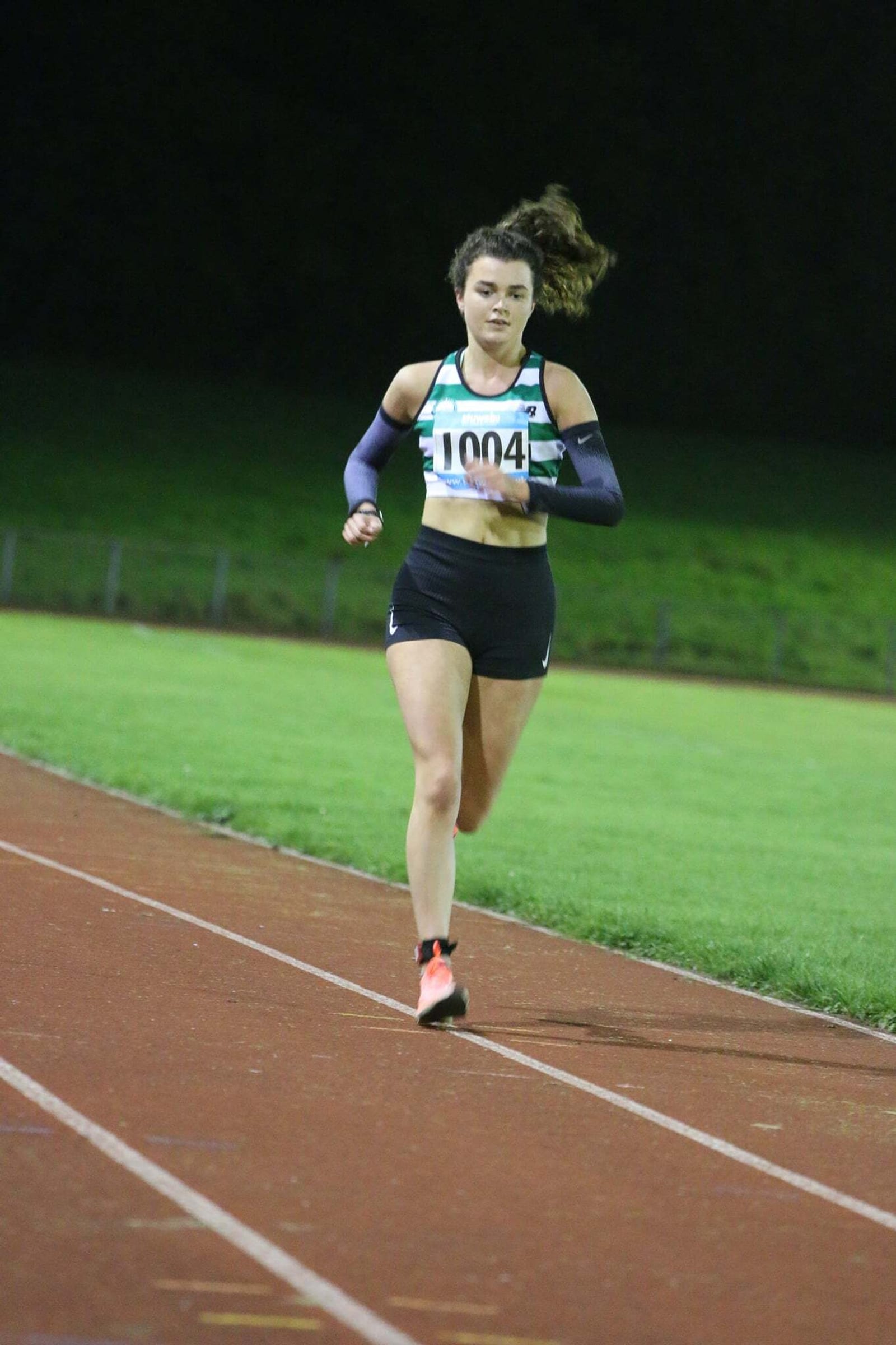 A runner with long hair in a ponytail competes on a track at night. She is wearing a green and white jersey, black shorts, and athletic shoes. Her bib number is 1004, and she appears focused as she runs on the red track with a grassy area in the background.