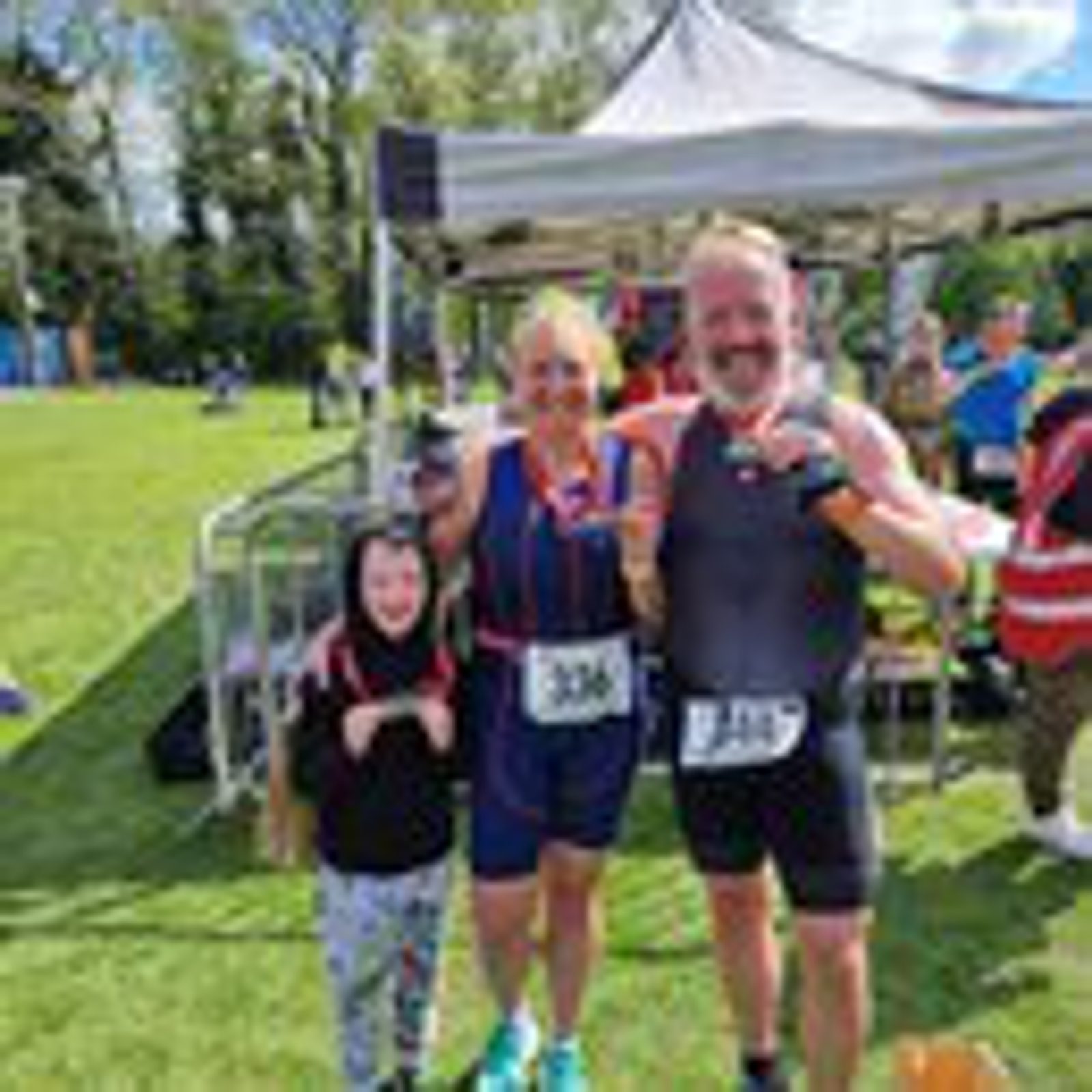 Three people stand together under a tent on a grassy field, smiling and holding medals. They are wearing athletic clothing and race bibs, indicating they have participated in an event. The background shows other participants and trees on a sunny day.