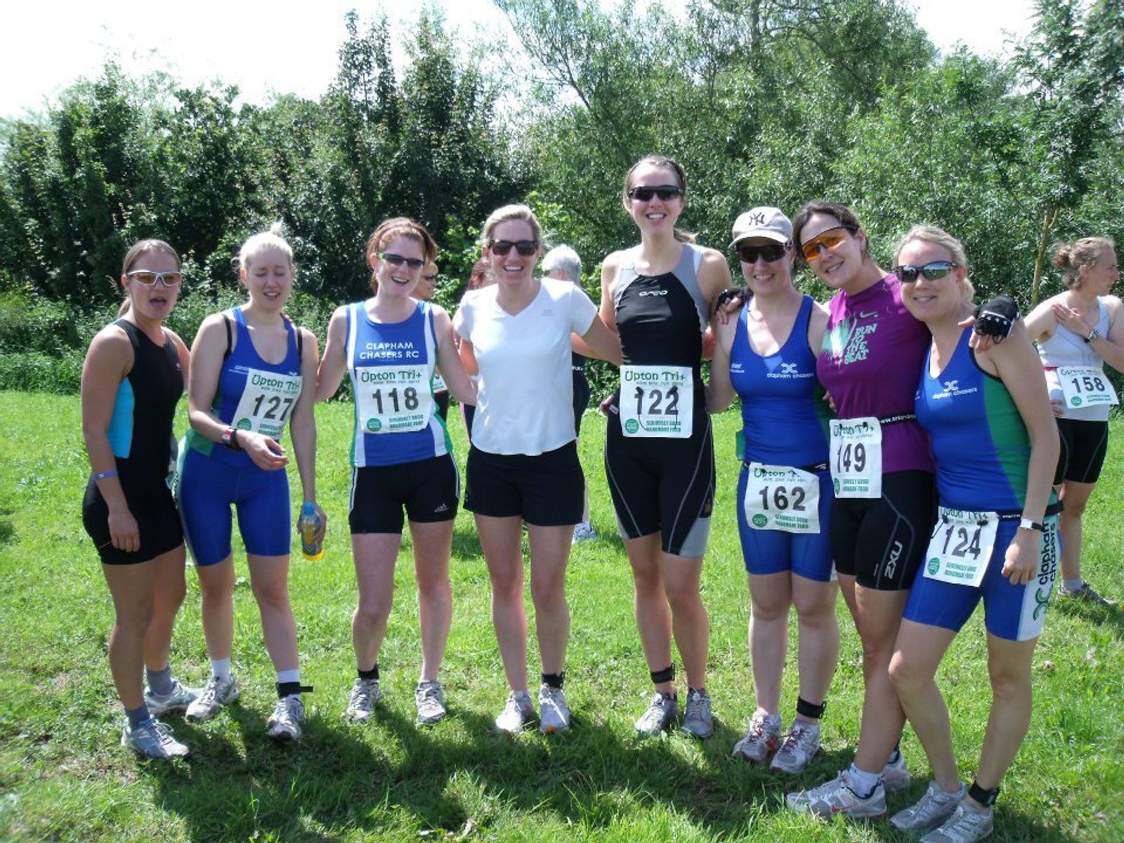 A group of eight women in athletic gear stand outdoors on grass, smiling for the camera. They each have race bibs pinned to their outfits, and some wear sunglasses. Trees and a clear sky are in the background, indicating a sunny day.