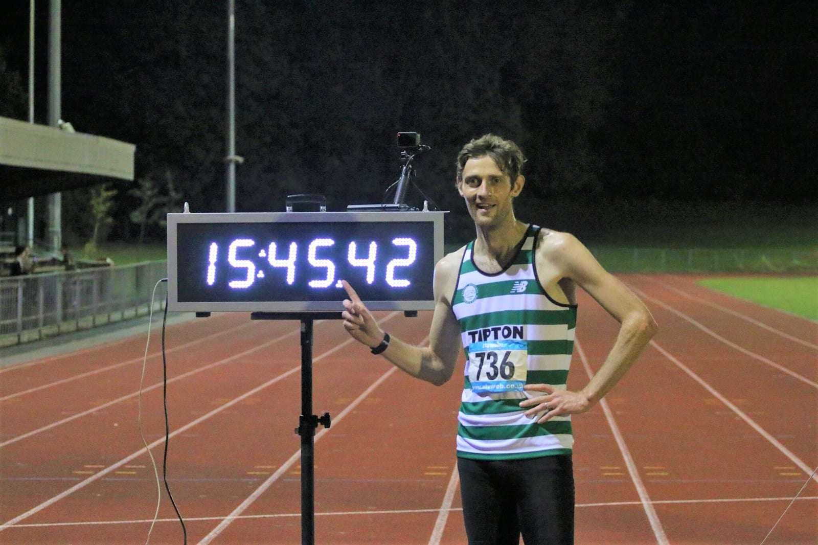 A runner in a green and white striped singlet and black shorts stands on a track at night, pointing to a digital timer displaying "15:45.42." The runner is smiling and has a race number 736 pinned to the singlet. The background shows a dimly lit athletic field.