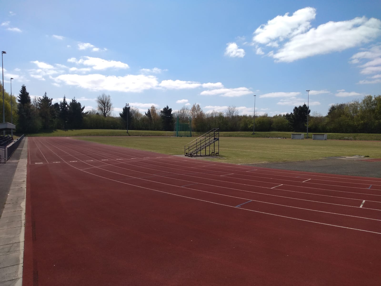 A photo of an empty outdoor athletics track on a sunny day. The red running lanes encircle a grassy field, with steps leading up to a small spectator stand. Trees and shrubs line the background under a blue sky with scattered white clouds.