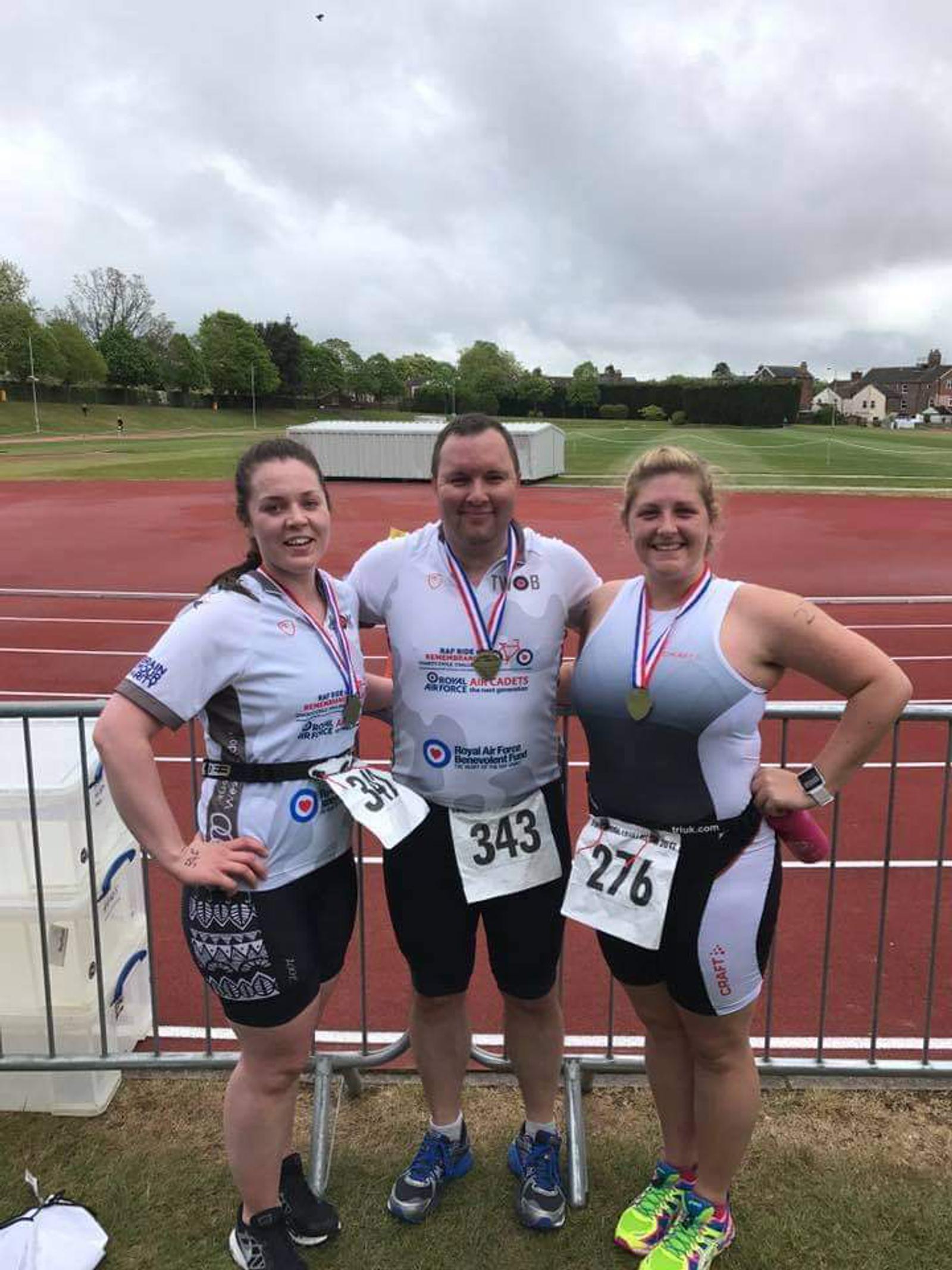 Three athletes wearing medals and numbered race bibs pose together after a race on an athletic track. They are all smiling and dressed in athletic gear. The background shows a grassy area and overcast sky.