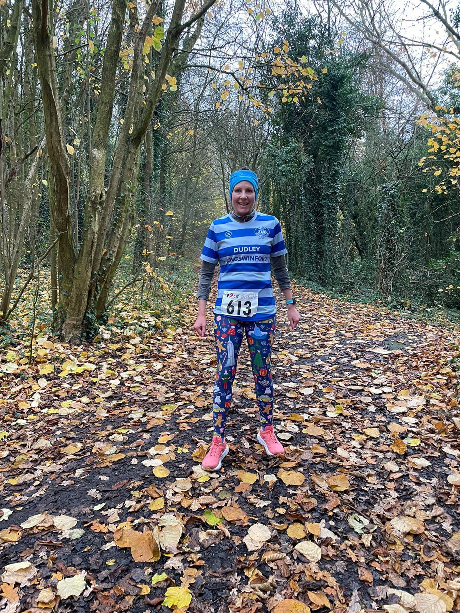 A runner stands on a leaf-covered path in a forest, wearing a blue striped shirt and colorful leggings. The person has a race bib with the number 613 and smiles at the camera. Trees and fallen leaves surround the scene.