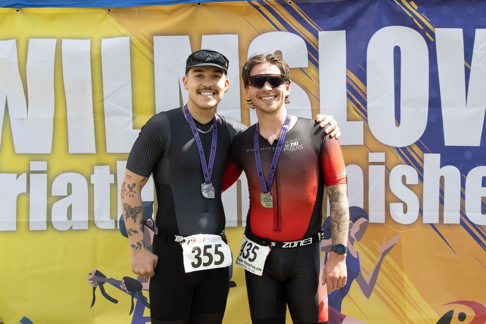 Two male triathletes wearing medals and race bibs (355 and 435) smile and pose in front of a colorful "Wilmslow Triathlon Finisher" banner, both dressed in athletic gear.