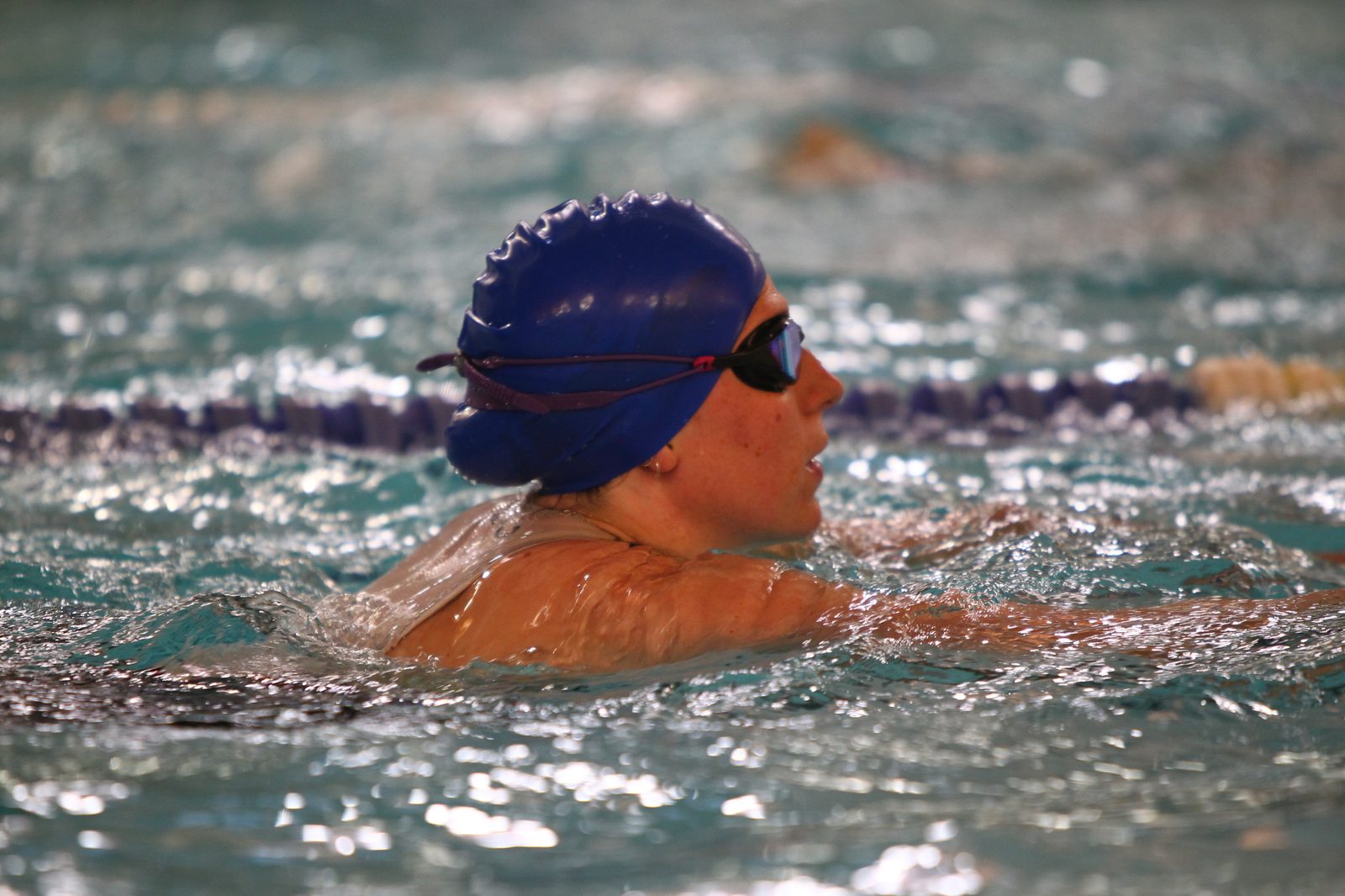 Person swimming in a pool wearing a blue swim cap and goggles. The swimmer is in profile view, with the water rippling around them as they move forward.