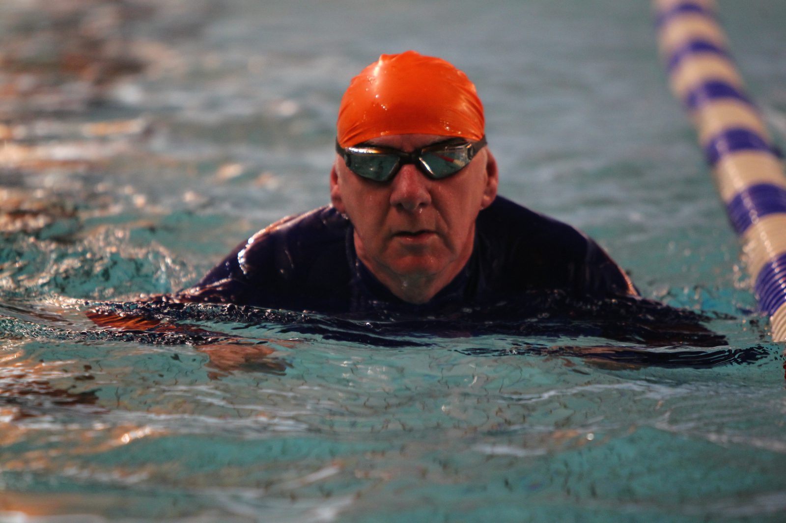 A swimmer wearing an orange swim cap and goggles is swimming in a pool, with their head above the water near a lane divider. The water is calm, and the lighting is dim.