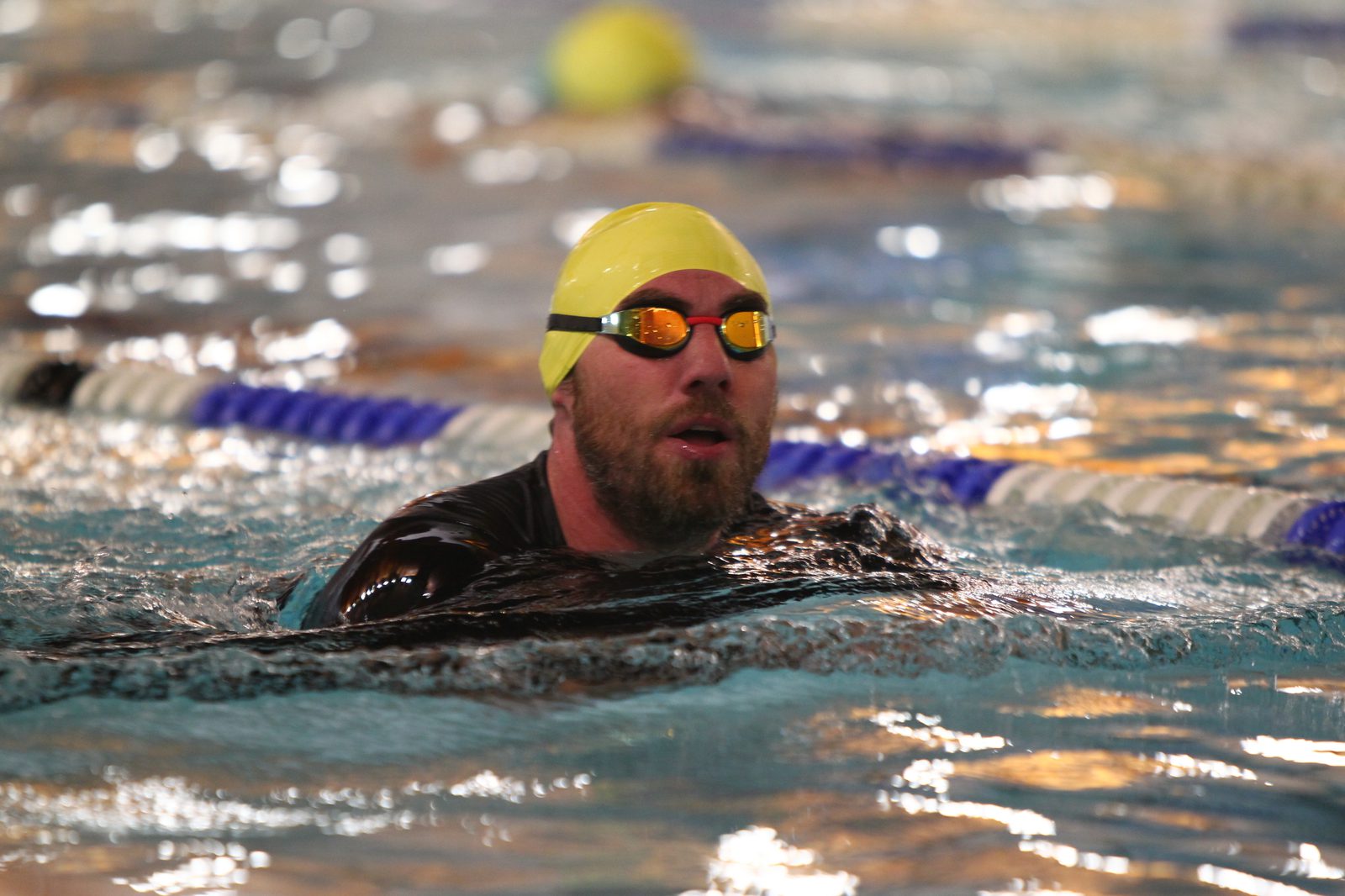 A swimmer with a beard and wearing a yellow swim cap and goggles swims through a pool. The pool lanes are marked by blue and white ropes, and the water is reflecting lights.