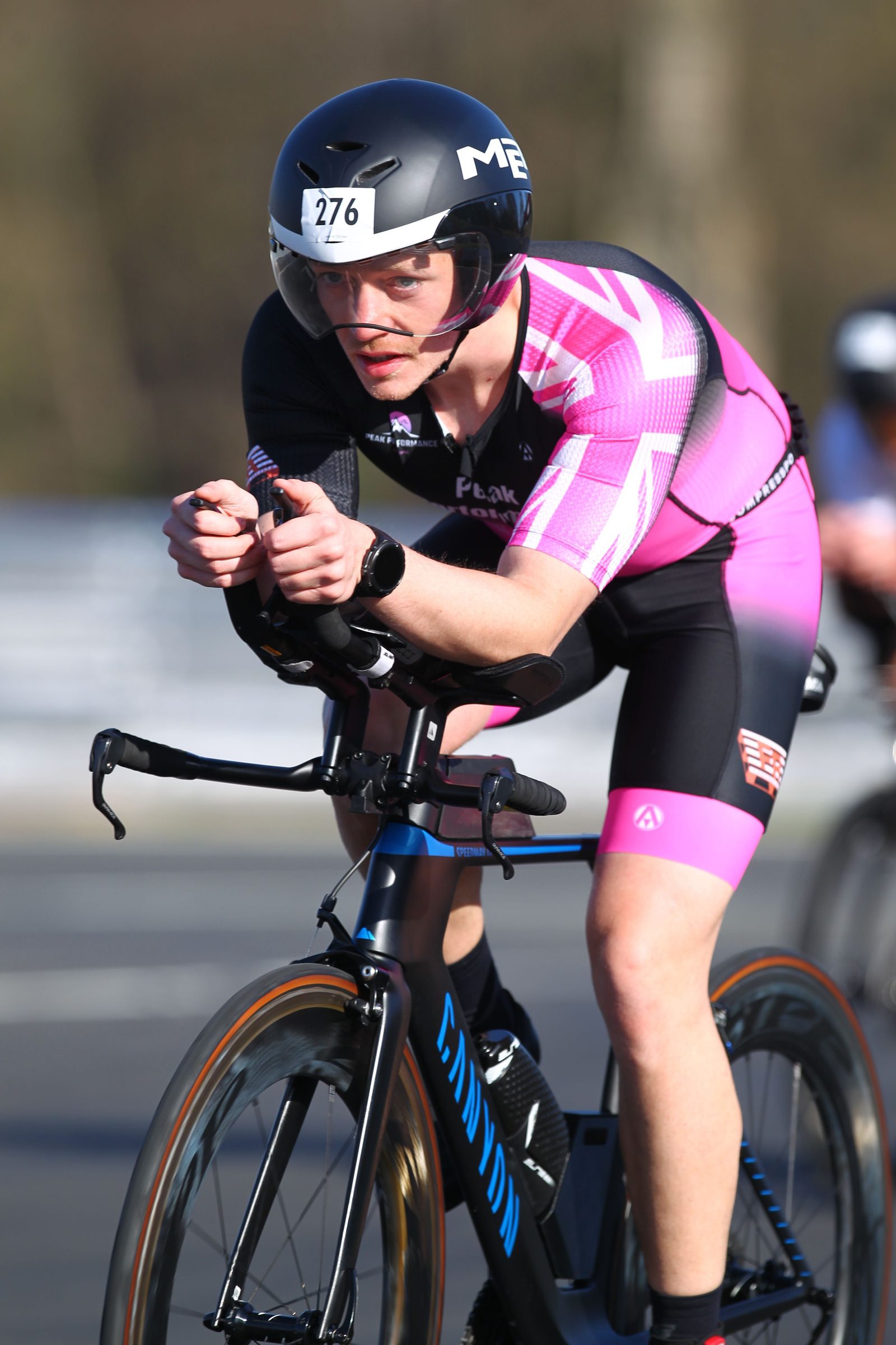 A cyclist wearing a pink and black racing suit and a black helmet labeled "276" rides a black Canyon bike in an aerodynamic position during a race. The background is blurred, emphasizing motion.