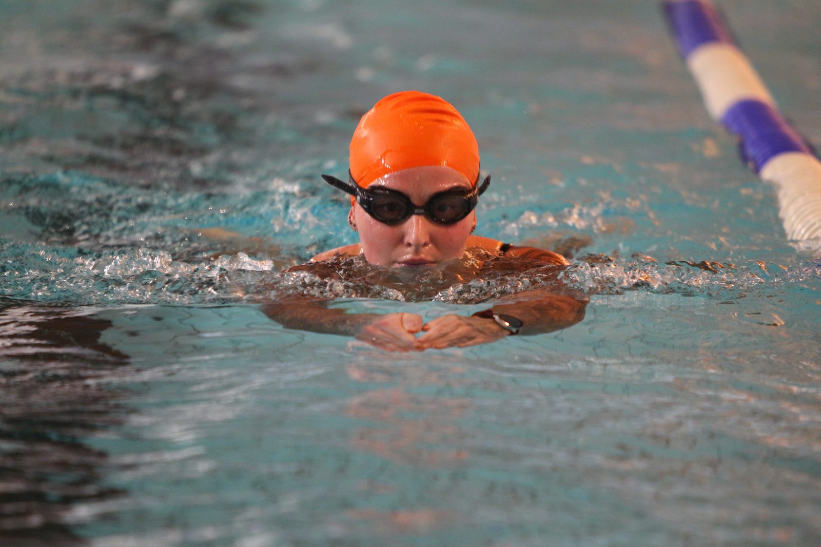 A swimmer wearing an orange cap and dark goggles practices breaststroke in an indoor pool, with a blue and white lane marker visible nearby.