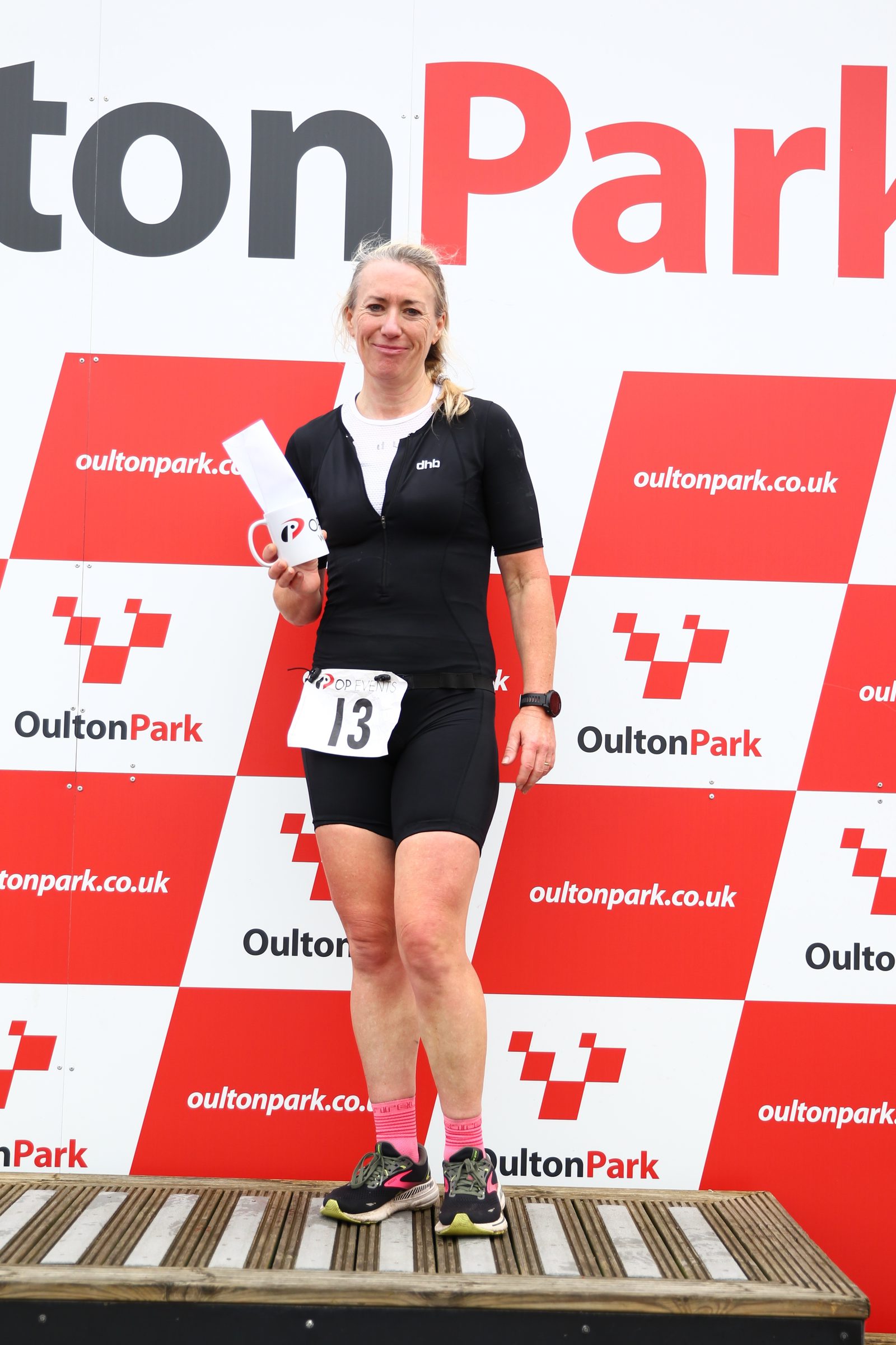 A woman in a black athletic outfit stands on a podium holding a mug and a rolled-up paper, smiling at the camera. The background features a red and white Oulton Park logo and checkered pattern.