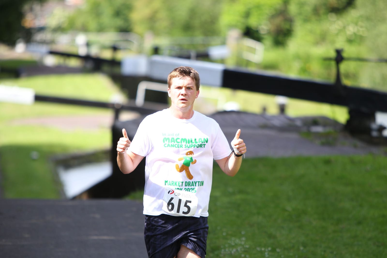 A man running outdoors gives two thumbs up. He wears a white "Macmillan Cancer Support" t-shirt and a race bib numbered 615. The background is green and sunny, with a blurred path and canal lock.