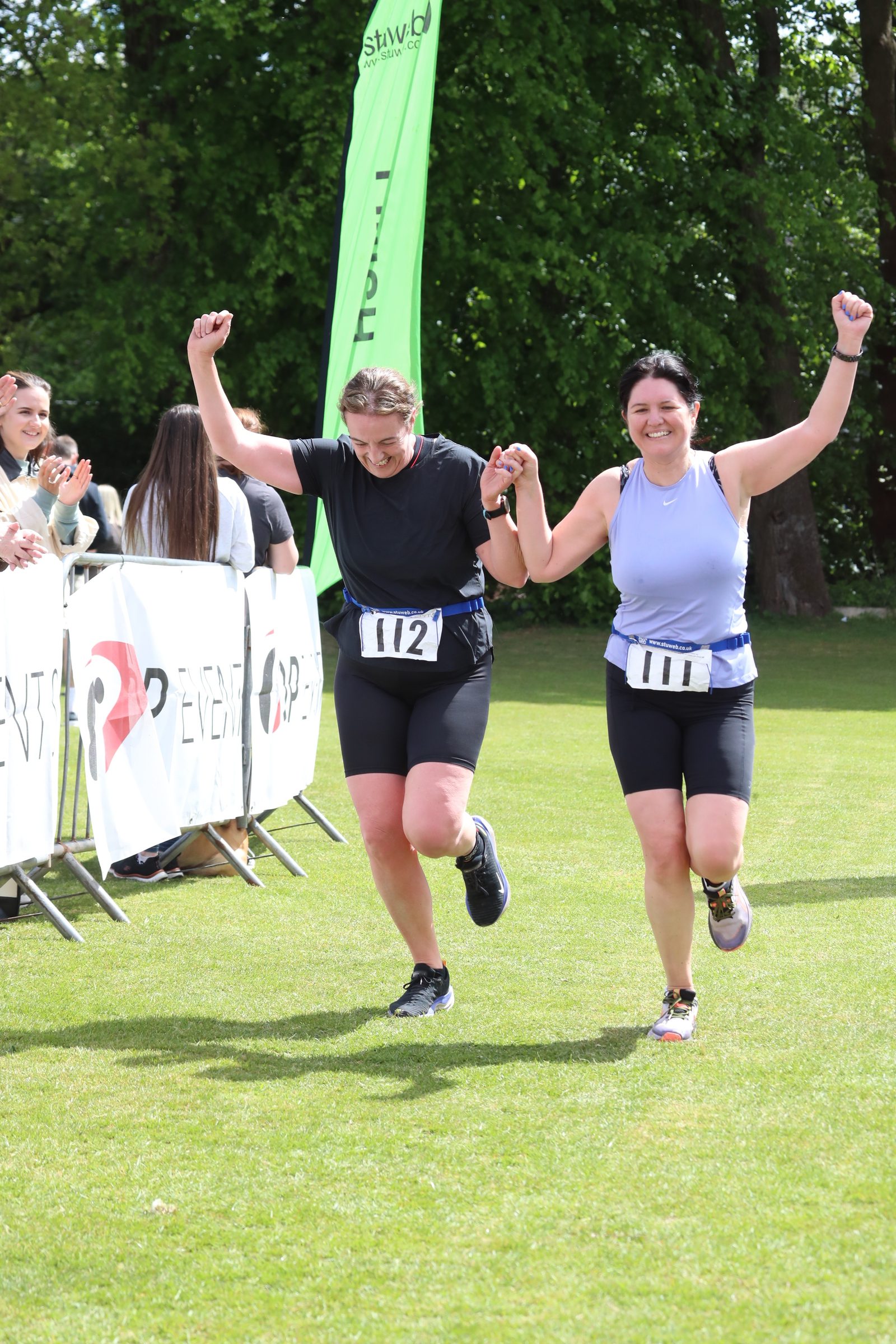 Two women holding hands and smiling as they cross the finish line of a race, both wearing athletic gear and numbered bibs (112 and 114), with cheering spectators and green trees in the background.