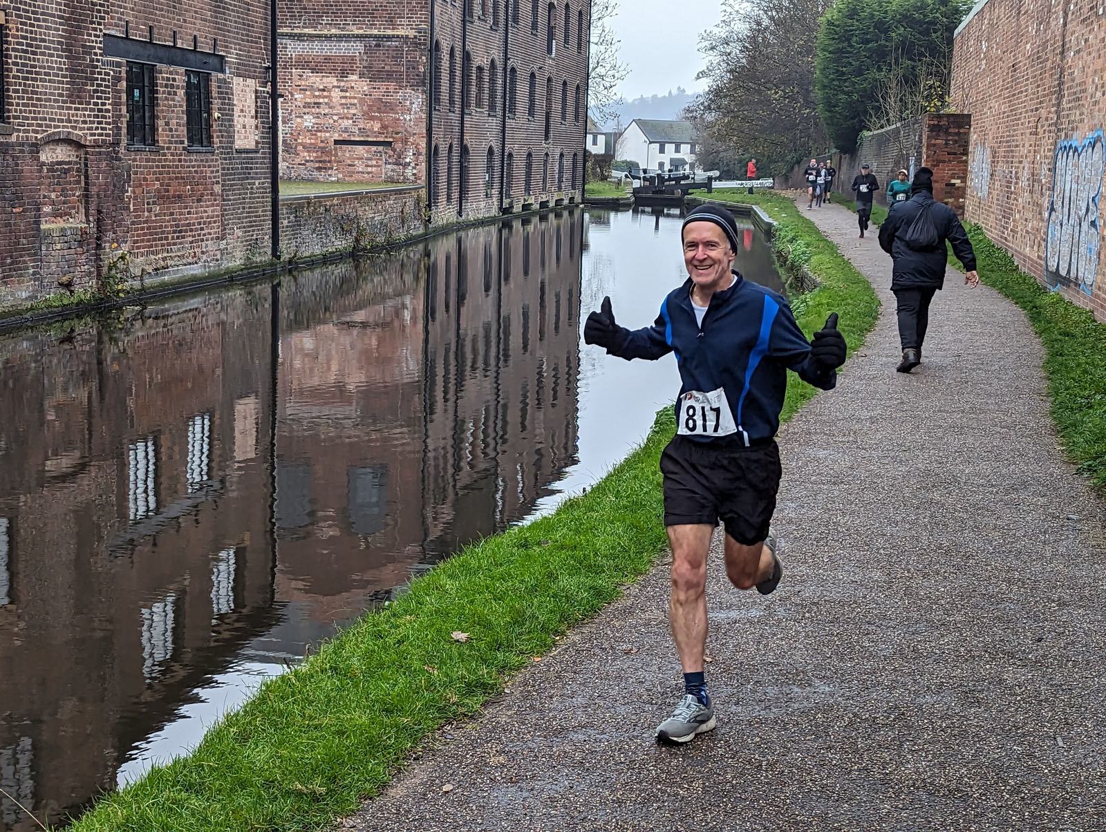 A runner in a blue and black outfit smiles and gestures with both thumbs up while jogging along a narrow canal path. The water reflects nearby buildings. Other people are walking and running in the background on an overcast day.