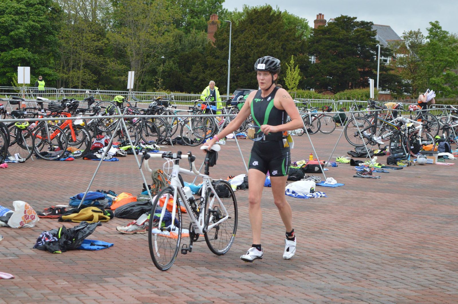 A triathlete in a black and green suit, wearing a helmet, walks with a white racing bicycle in a transition area filled with other bikes and scattered gear. The background shows a paved surface, trees, and additional athletes preparing for their next stage.