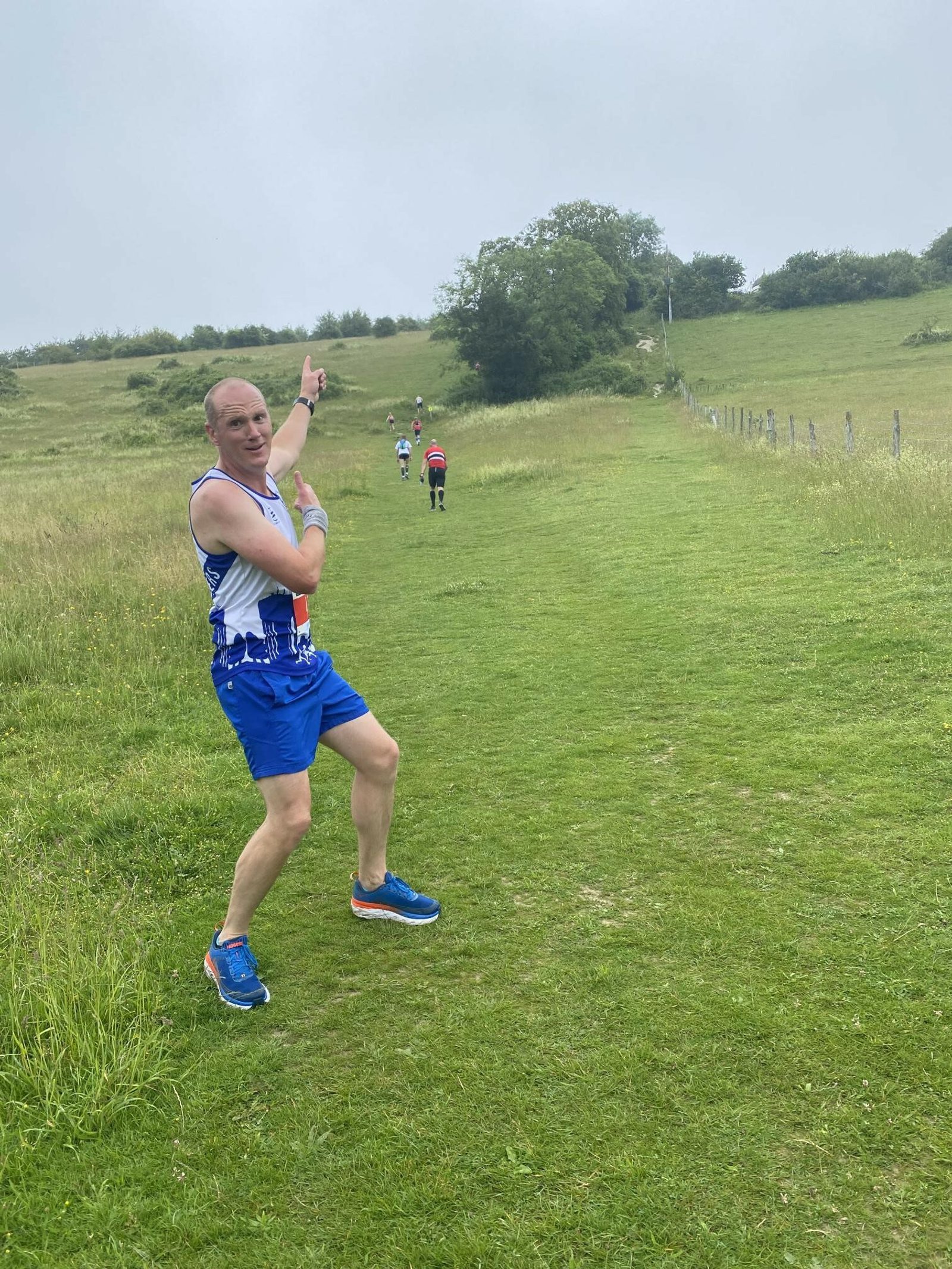 A person in blue athletic gear, including a white and blue tank top and shorts, poses playfully while pointing towards a group of runners ahead on a grassy trail. The scene is set against a hilly background with overcast skies.