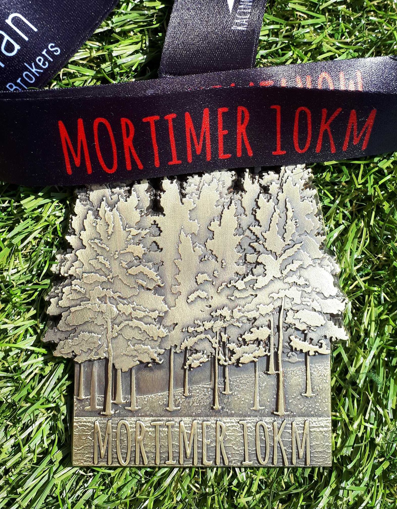 A silver medal with an engraved design of trees stands on green grass. The medal is attached to a black and red lanyard with the text "MORTIMER 10KM." The medal also has the words "MORTIMER 10KM" inscribed at the bottom.