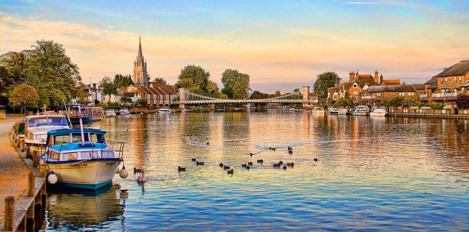 A picturesque river scene at sunset with parked boats along the left side, ducks swimming in the water, and a suspension bridge leading to a quaint village with a prominent church tower and houses surrounded by trees in the background.