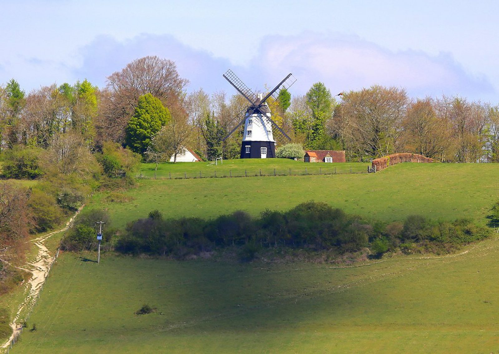 A traditional white and black windmill stands atop a lush, green hill surrounded by trees with fresh spring foliage. A narrow dirt path winds up the hill to the windmill. In the background, there's a small cottage and a wooden fence. The sky is clear with light clouds.