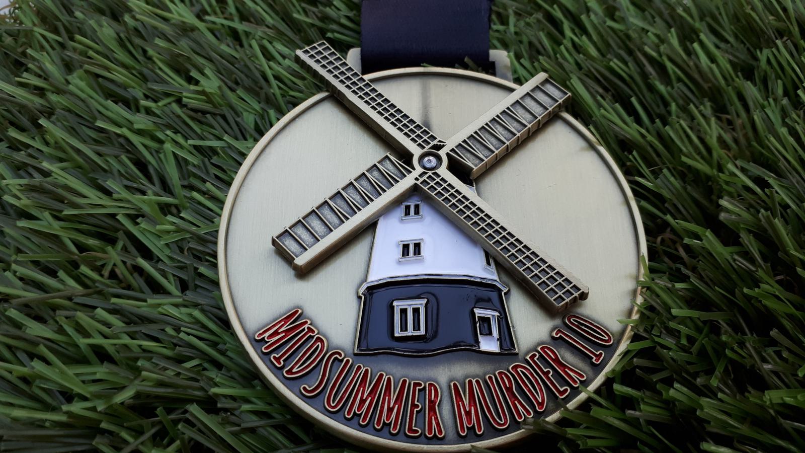 A circular medal with a windmill design in the center, reading "MIDSUMMER MURDER 10" at the bottom. The medal is placed on a background of artificial green grass, and a black ribbon is attached to the top of the medal.