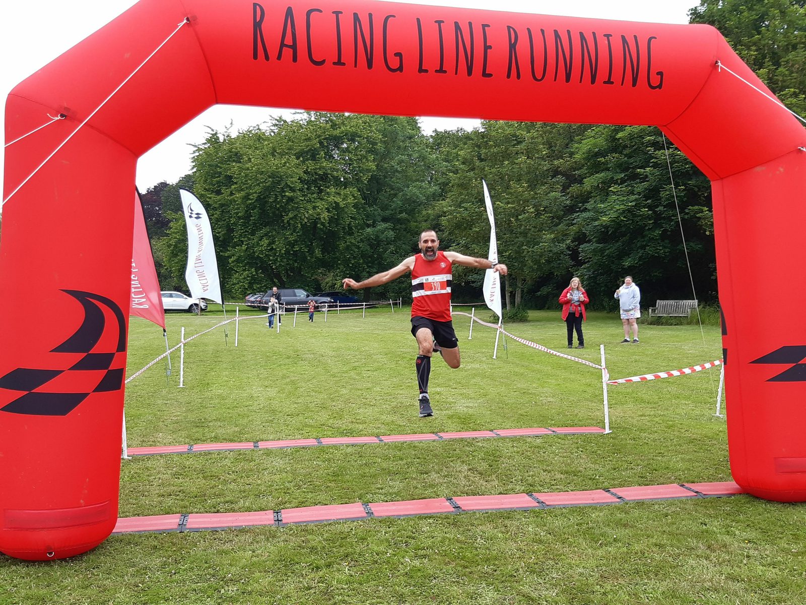 A man in a red and black running outfit is jumping as he crosses the finish line beneath a red inflatable arch that reads "Racing Line Running." The background is a grassy area surrounded by trees, with a few people standing nearby.