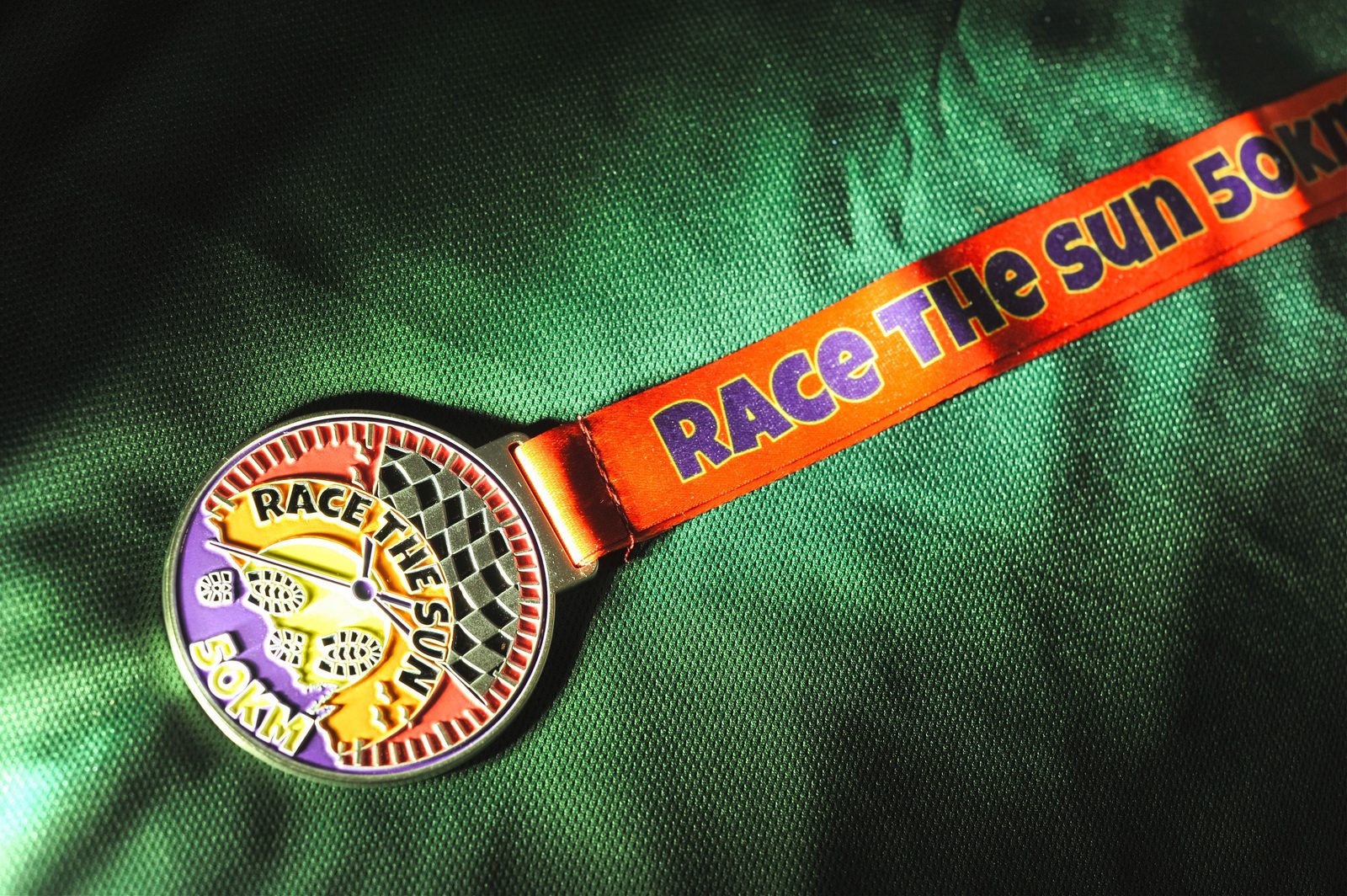 A colorful medal on a green fabric backdrop. The medal has an image of a setting sun and the text "RACE THE SUN 50 KM". The attached ribbon is orange and purple with the text "RACE THE SUN 50 KM" printed on it.