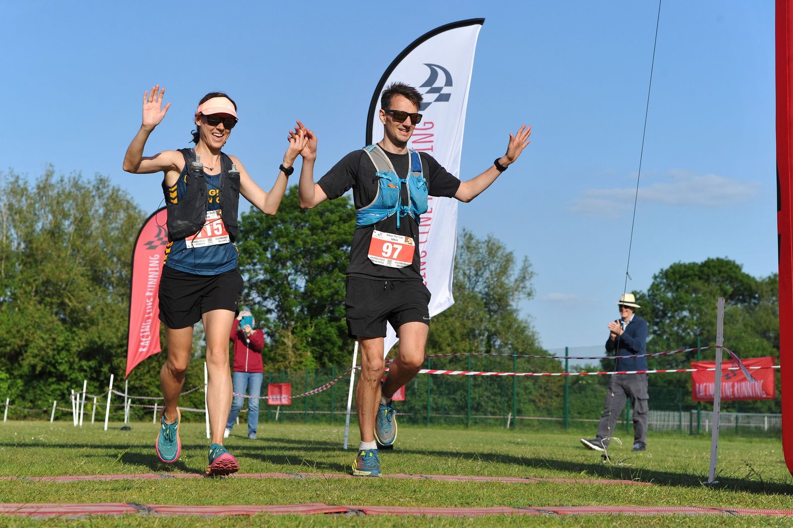 Two runners, a man and a woman, are crossing a race finish line on a sunny day, holding hands and raising their other hand in celebration. Both wear running gear, bib numbers, and sunglasses. There's a cheering crowd, race banners, and a clear blue sky in the background.